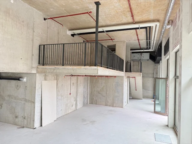 Interieur van een onafgewerkte ruimte met betonnen wanden, een verhoogd platform met zwarte railing, en zichtbare leidingen.