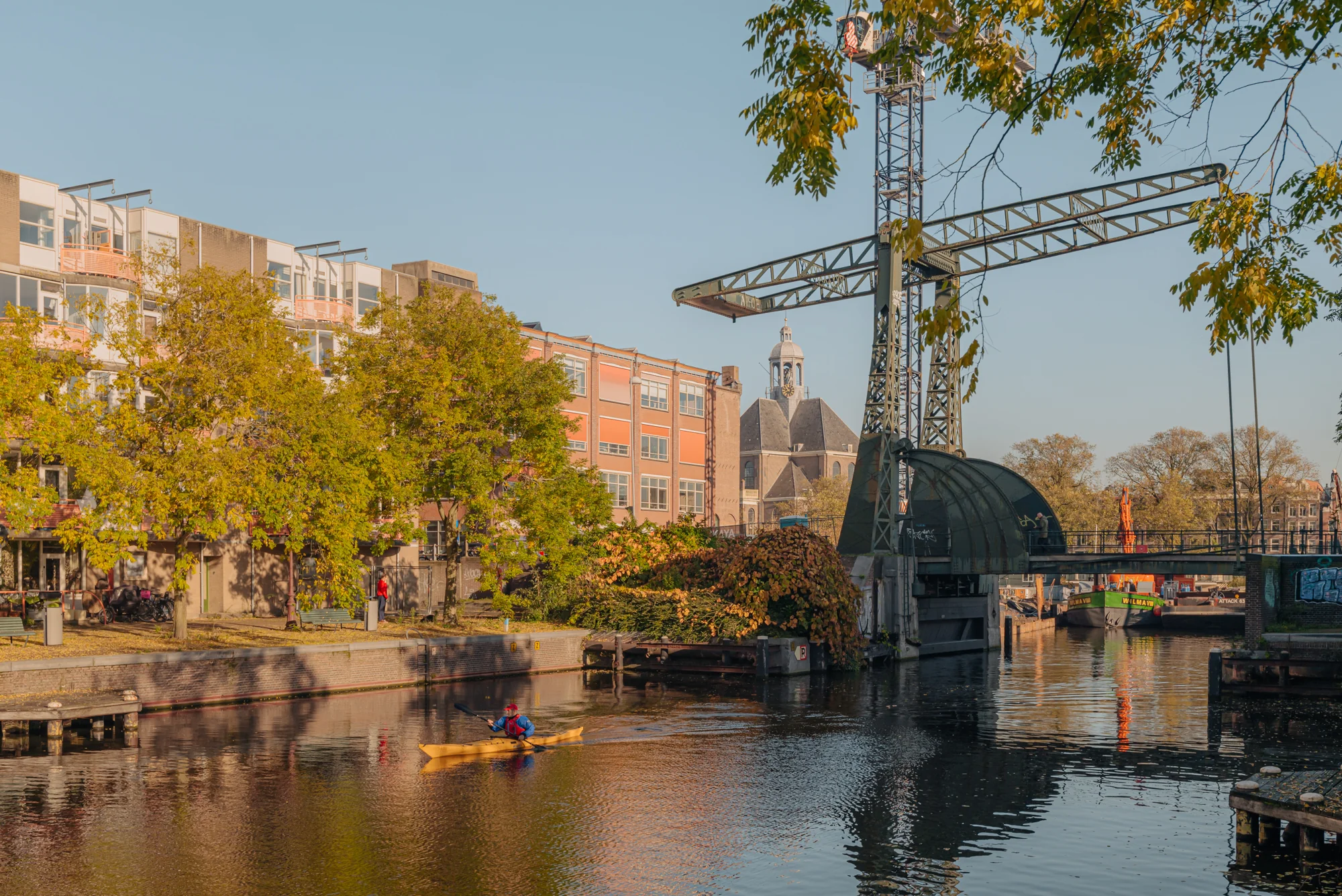 Een kajakker peddelt langs een gracht in de buurt van Hoogte Kadijk, omgeven door gebouwen en een oude draaibrug.