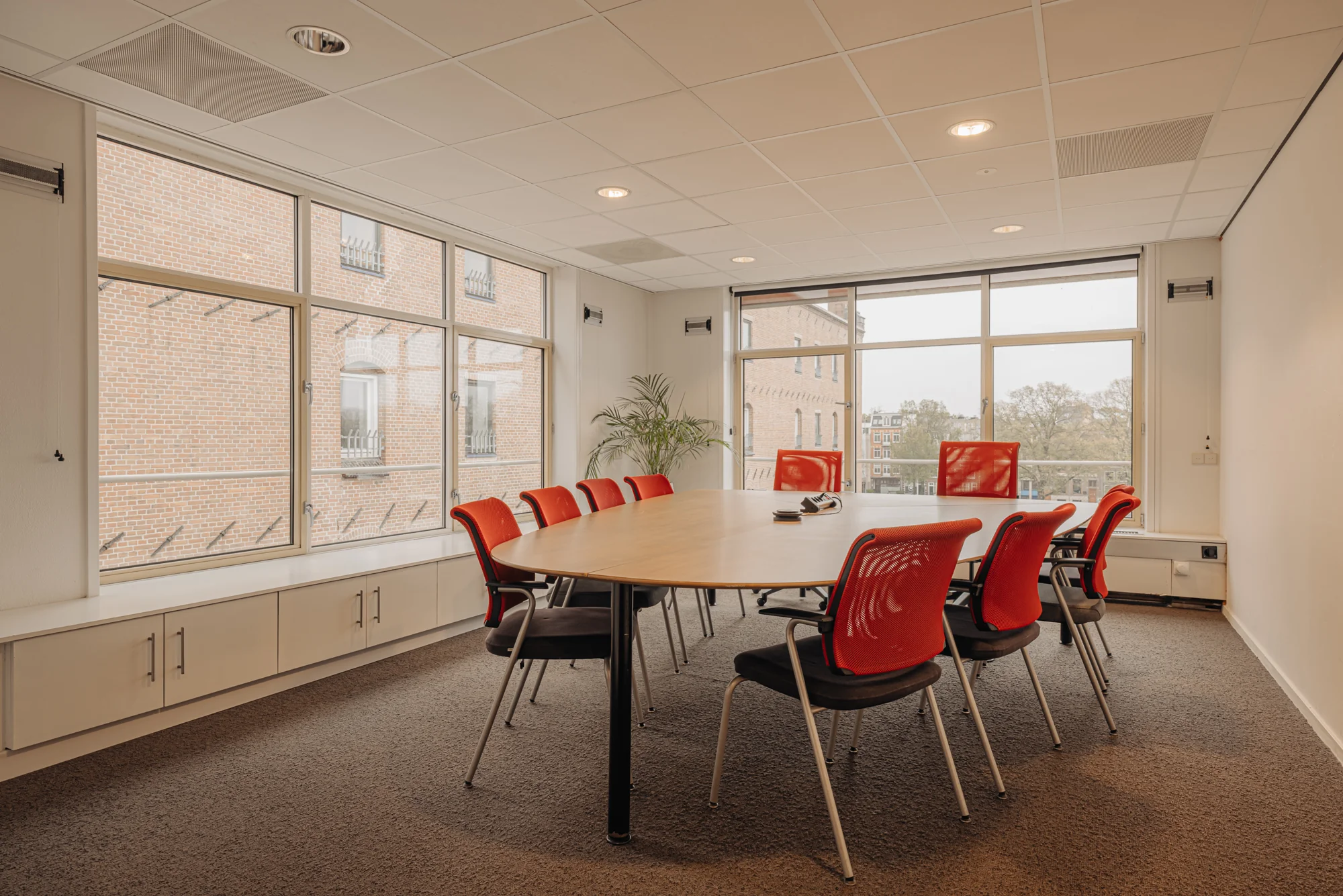 Vergaderruimte met een ovale tafel en rode stoelen bij een groot raam met uitzicht op een bakstenen gebouw.