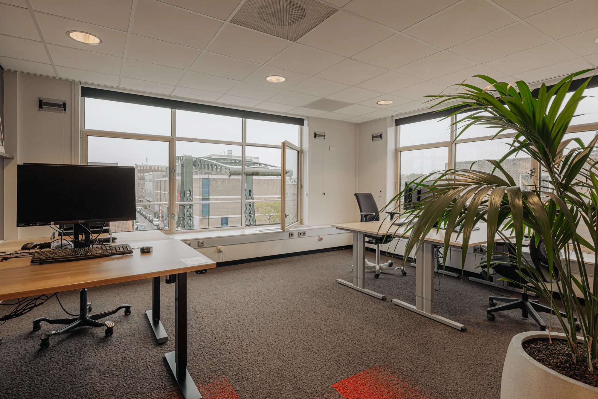 Een modern, leeg kantoor bij Hoogte Kadijk met bureaus, bureaustoelen en uitzicht op een gebouw buiten.