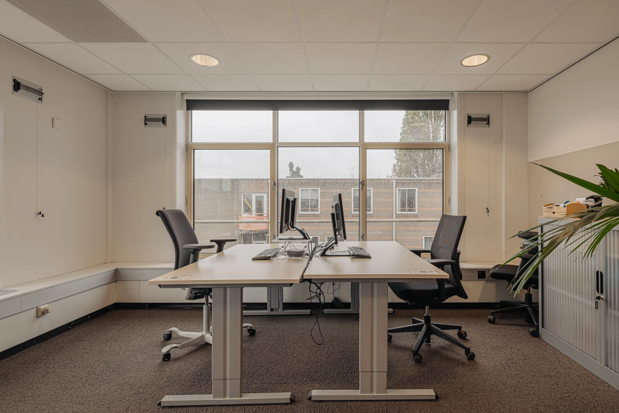 Een moderne kantoorruimte met twee werkplekken, bureaustoelen en computers, met uitzicht op de Hoogte Kadijk.