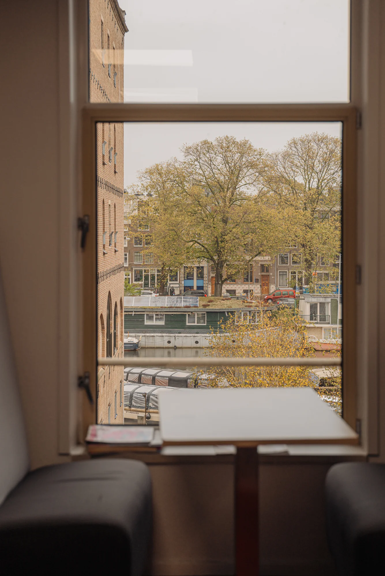Uitzicht vanuit het raam op de Hoogte Kadijk met bomen, gebouwen en een woonboot op de voorgrond.