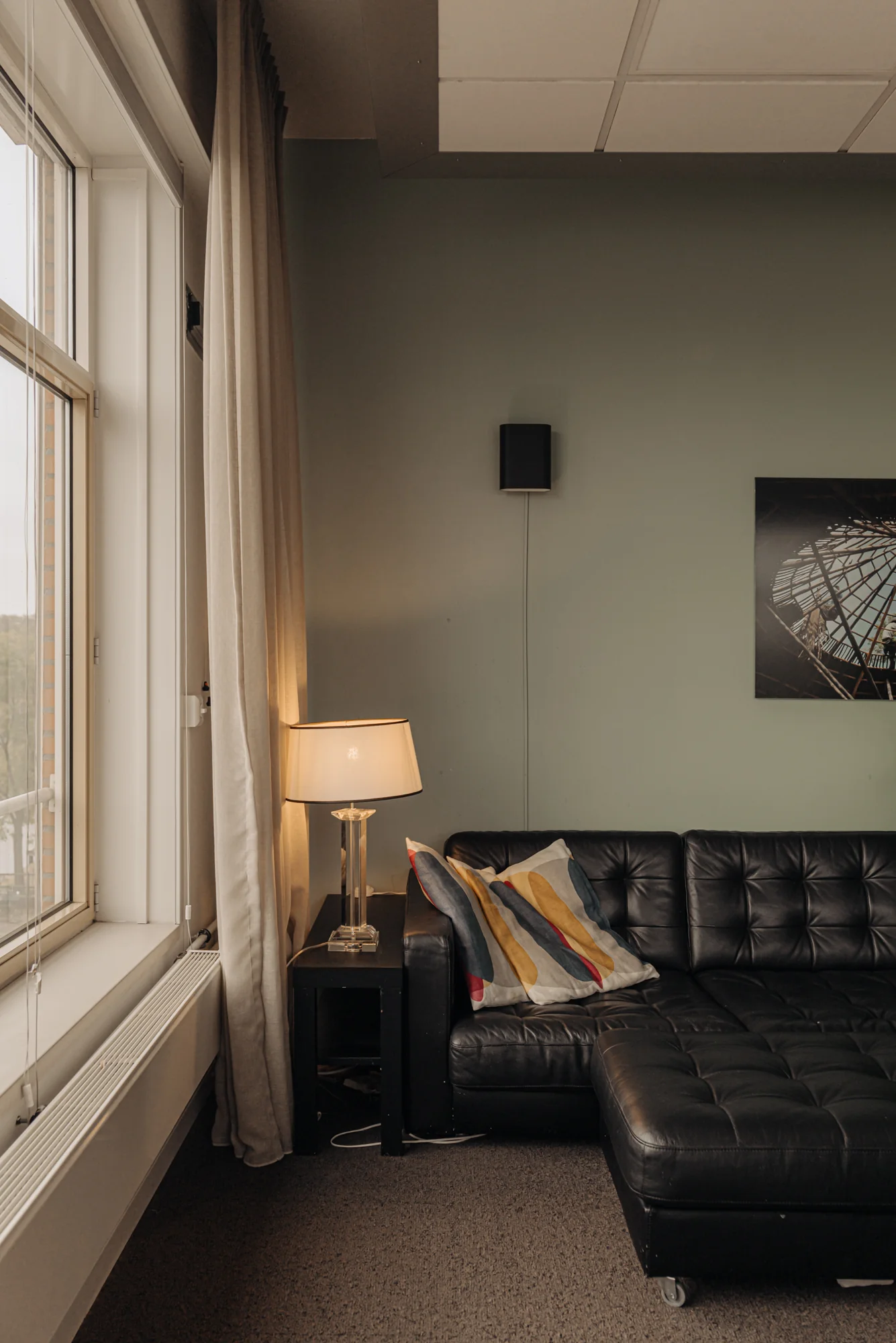 Een moderne woonkamer met een zwart leren bank, kleurrijk kussen, staande lamp en groot raam.