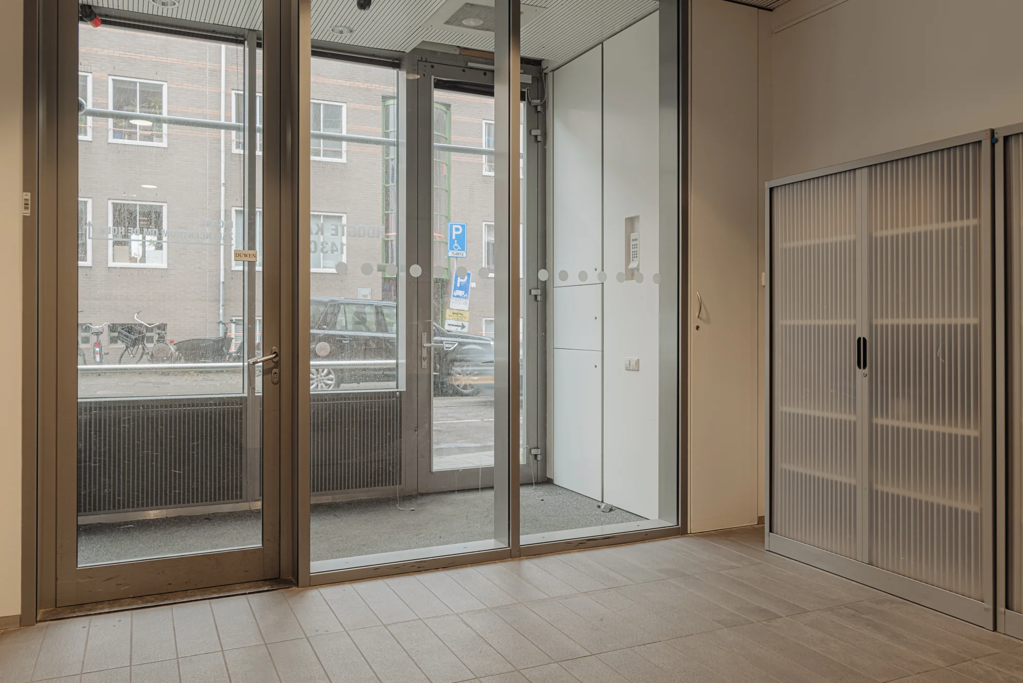 Een entree met glazen deuren en een (rol)kast, waar je doorheen de straat aan de Hoogte Kadijk kan zien.