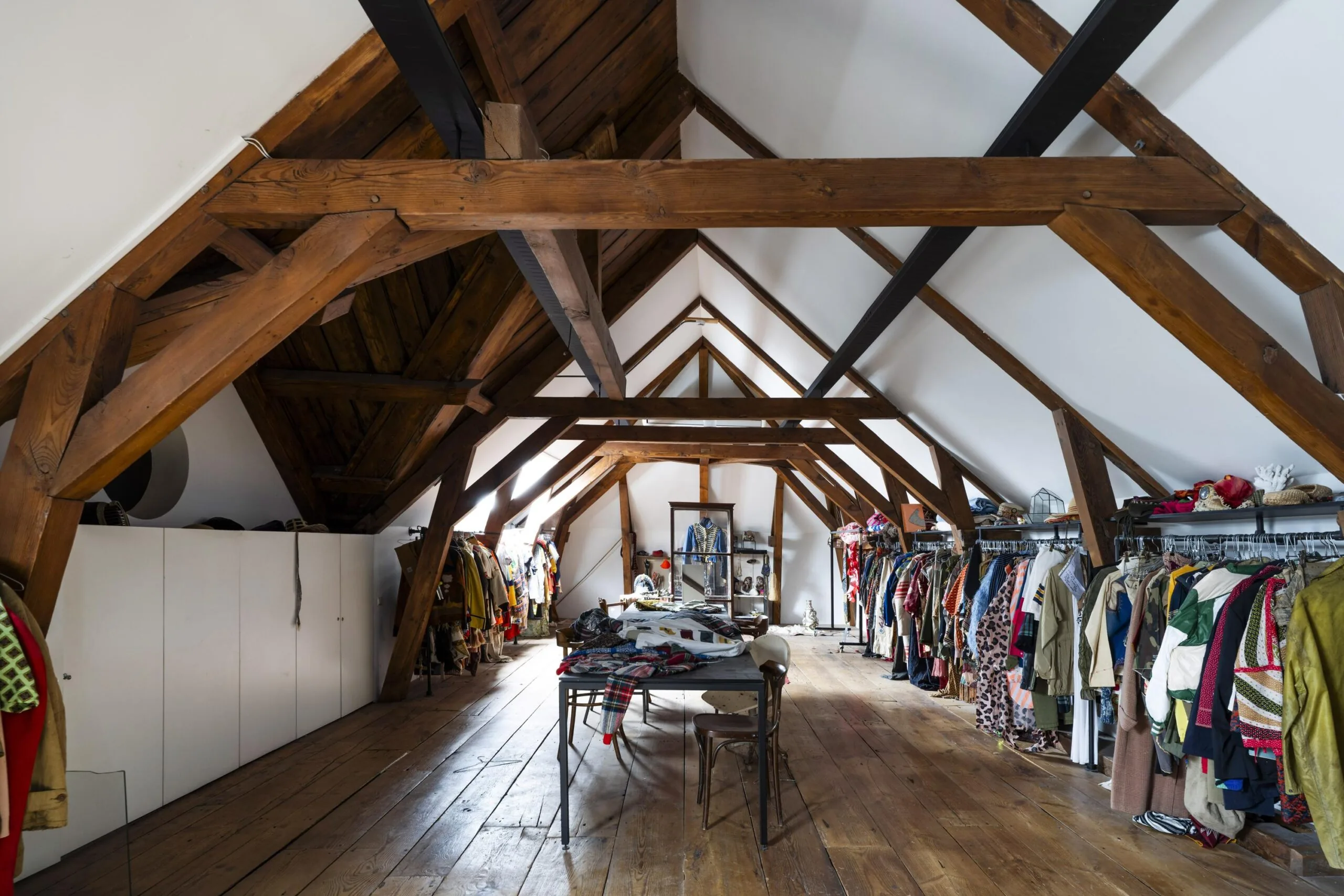 Een zolderruimte met houten balken, ingericht als kledingwinkel met rekken vol kleurrijke kleding.