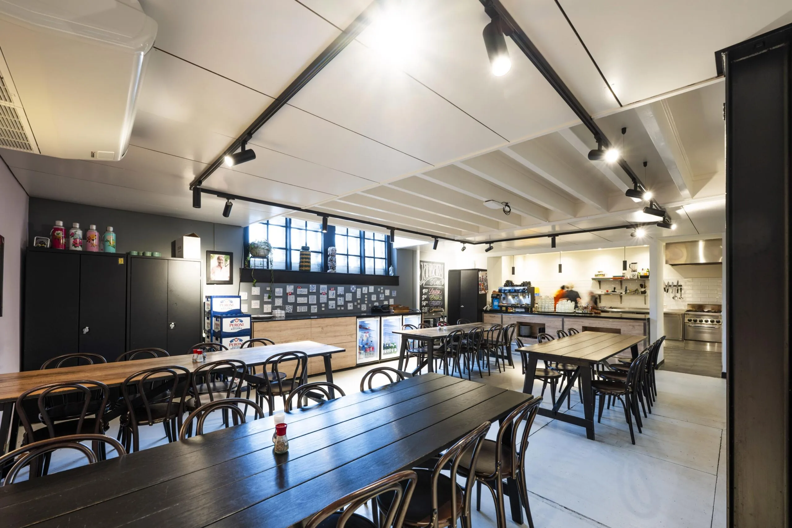Een ruime, moderne kantine met lange tafels en stoelen, verlichting, kasten en een keuken in de hoek.