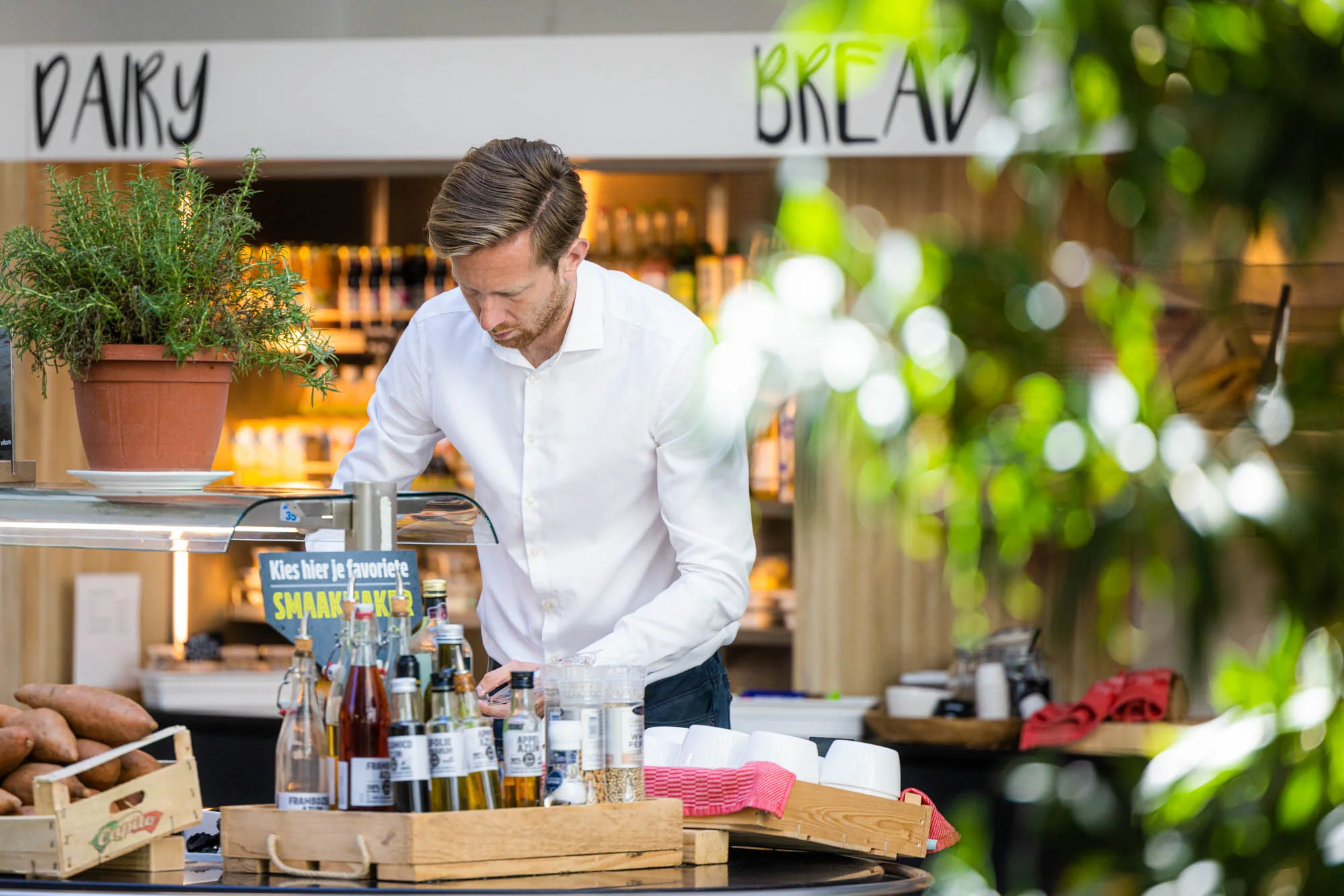 Een man in een wit overhemd werkt geconcentreerd aan een tafel met diverse flessen en kruiden, omgeven door groen in een winkelomgeving.