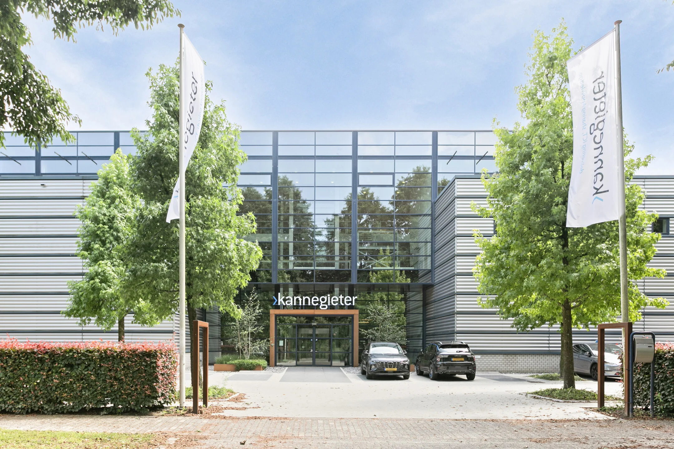 De ingang van het moderne Kannegieter-gebouw met vlaggen en auto's op de oprit.