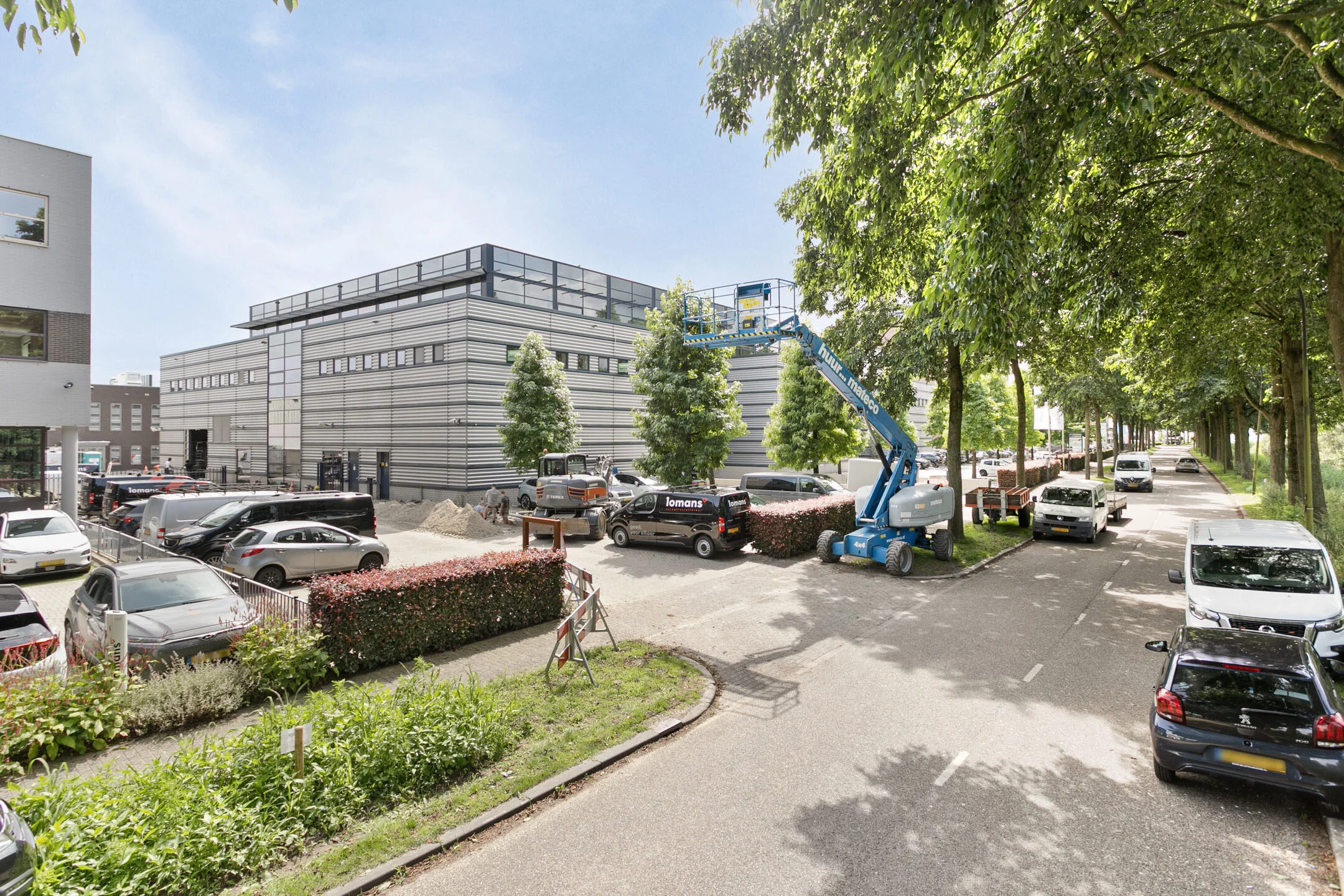 Een industrieel gebouw met een parkeerplaats en een blauwe hoogwerker aan de straatkant, omgeven door bomen en auto's.