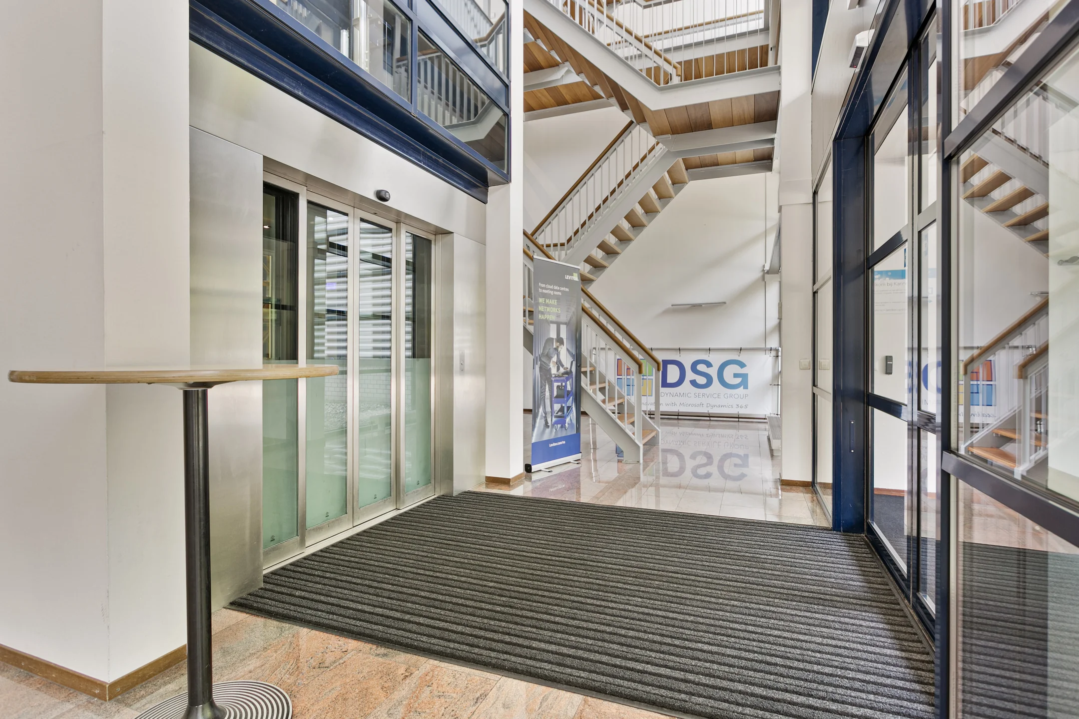 Ingang van een modern gebouw met een lift en trap, met een bord van Dynamic Service Group.