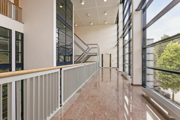Een moderne gang met glazen wand en trap in een kantoorgebouw.