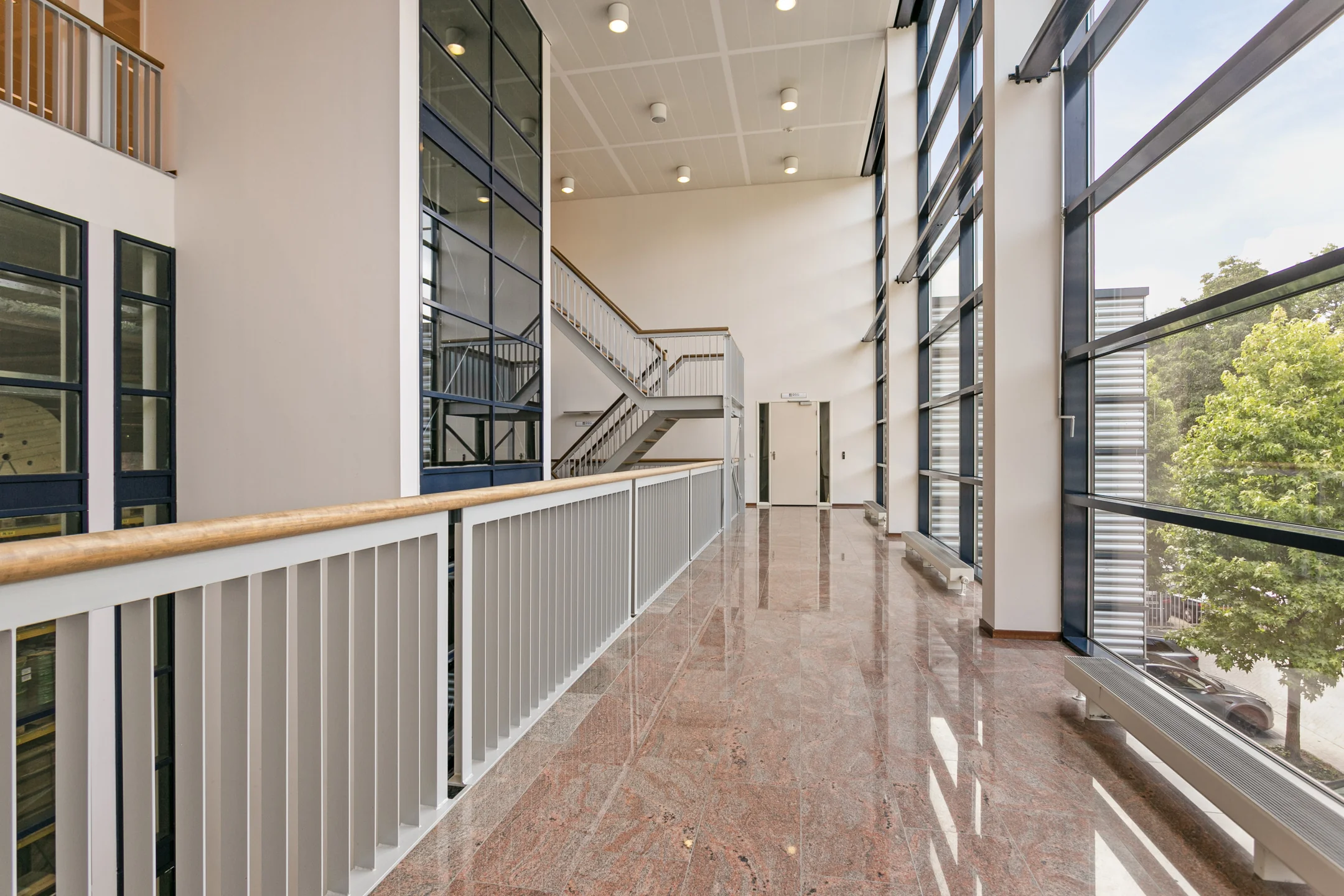Een moderne gang met glazen wand en trap in een kantoorgebouw.