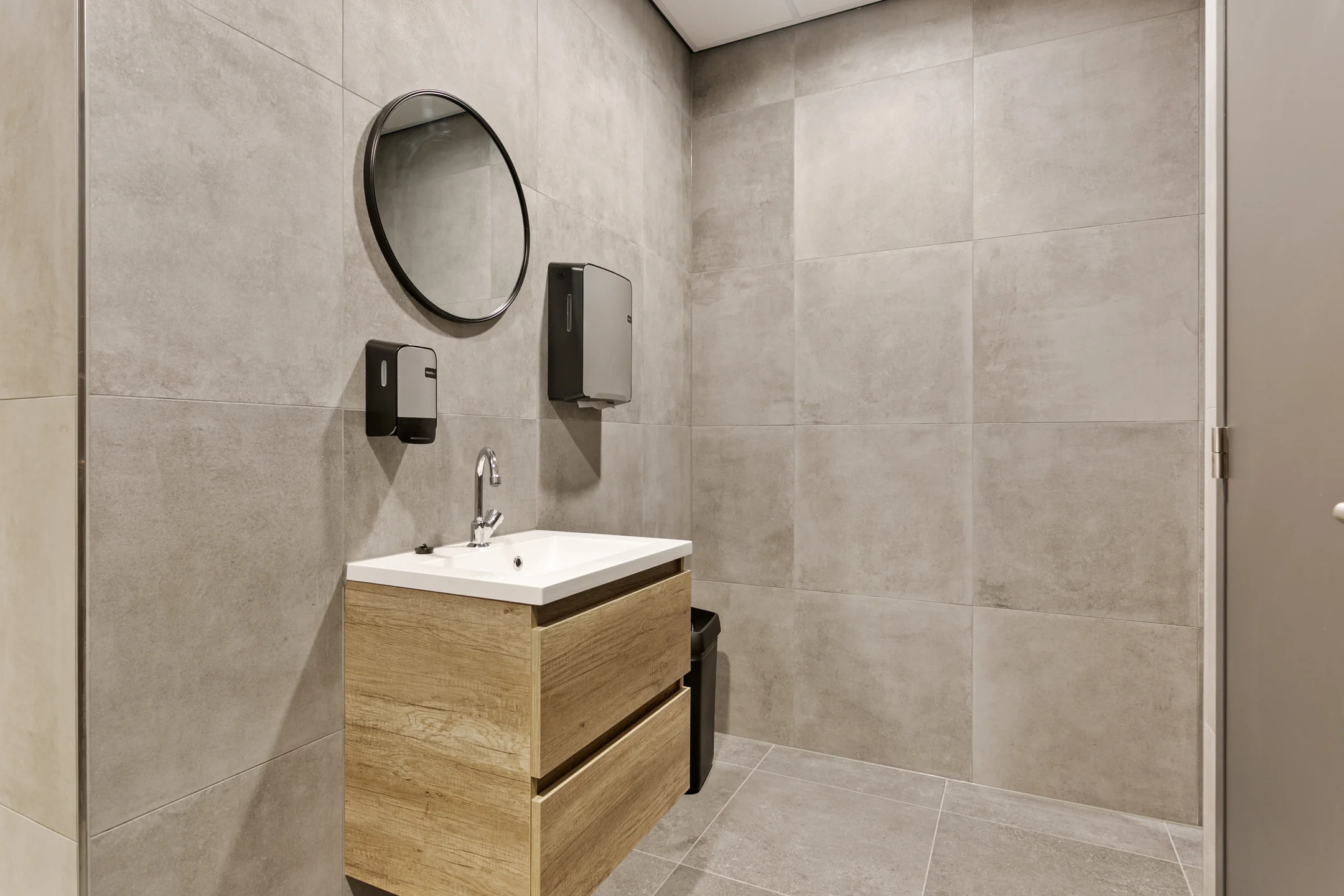 Een modern ingerichte badkamer met een houten wastafelmeubel, ronde spiegel, en grijze tegels.