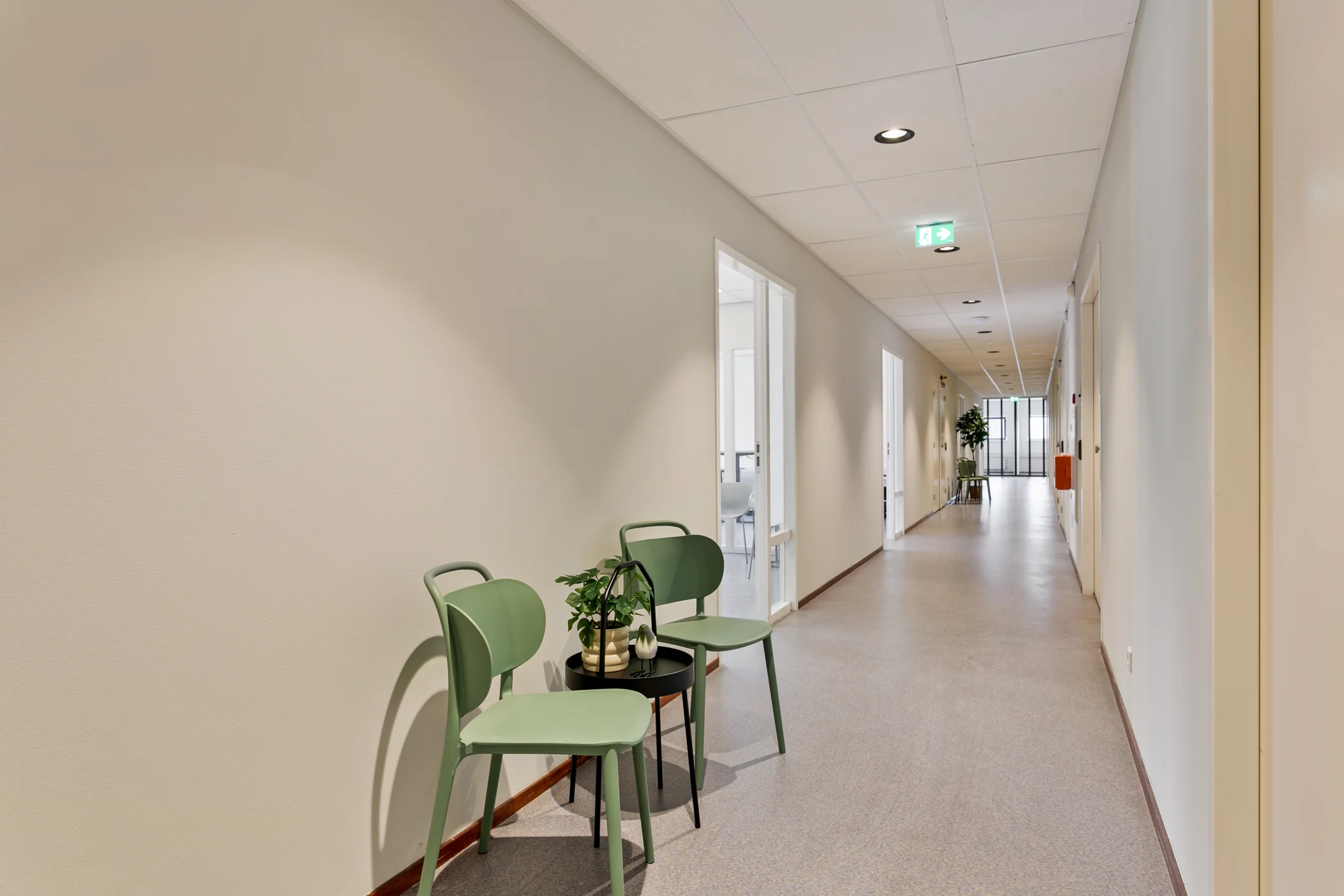 Een lange gang met groene stoelen en een tafeltje met planten tegen de muur.