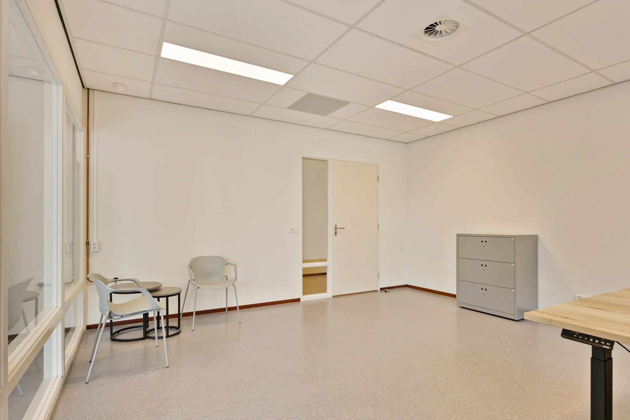 Een lege kantoorruimte met witte muren, twee stoelen, een kleine tafel, en een grijze ladekast.