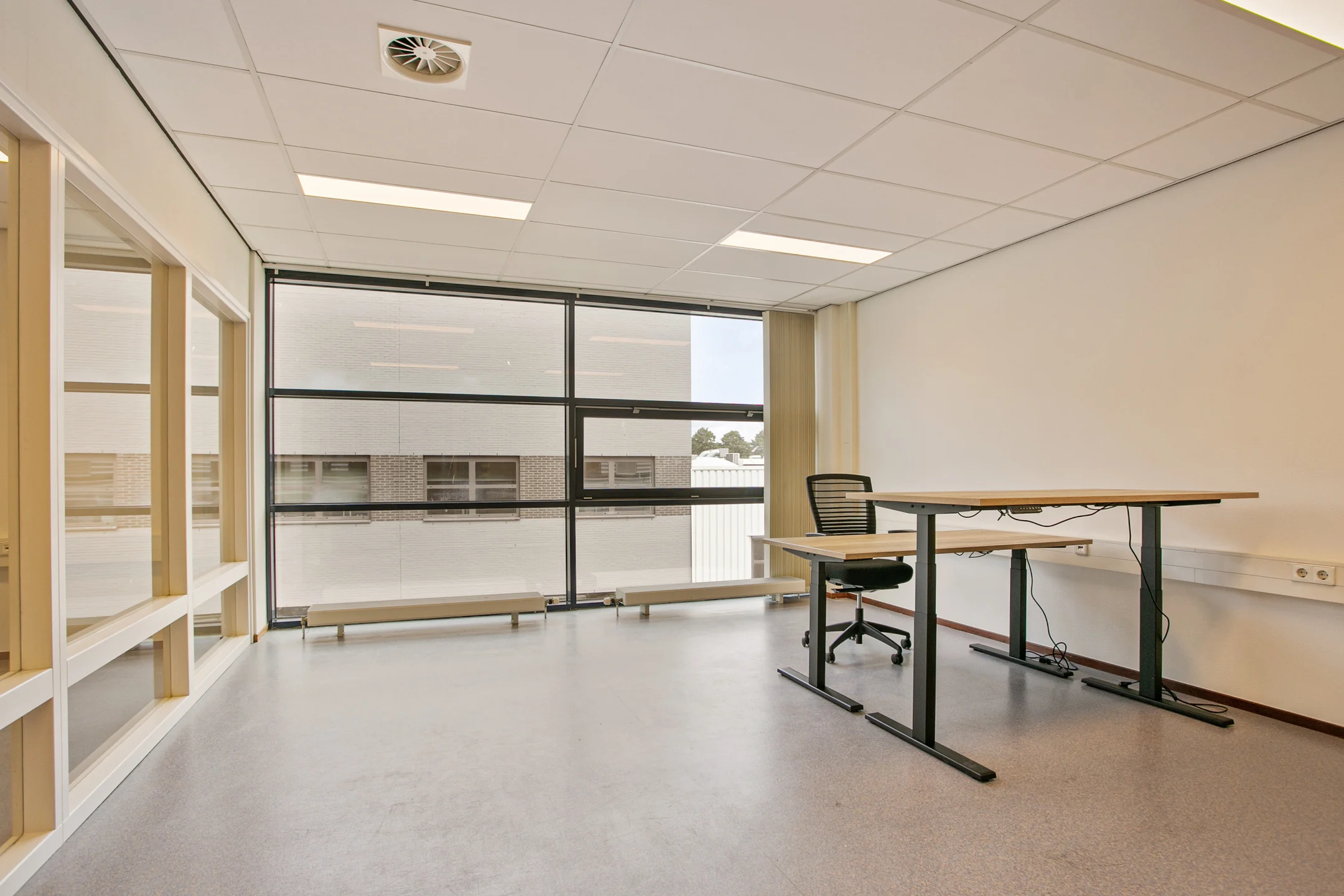 Een lege kantoorruimte met een bureau, bureaustoel en groot raam met uitzicht naar buiten.