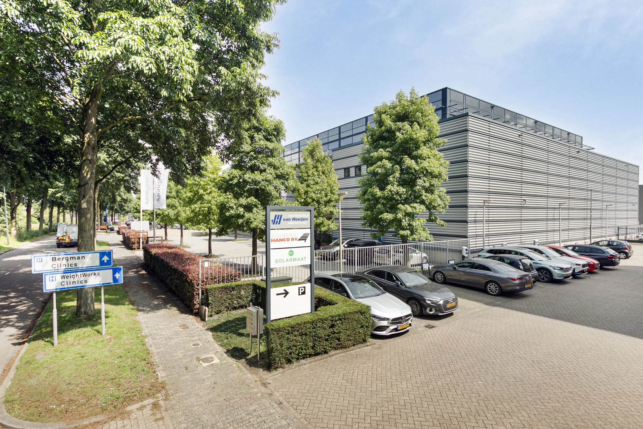 Parkeerplaats en bedrijfsgebouwen omringd door bomen, met bewegwijzering naar nabijgelegen klinieken.