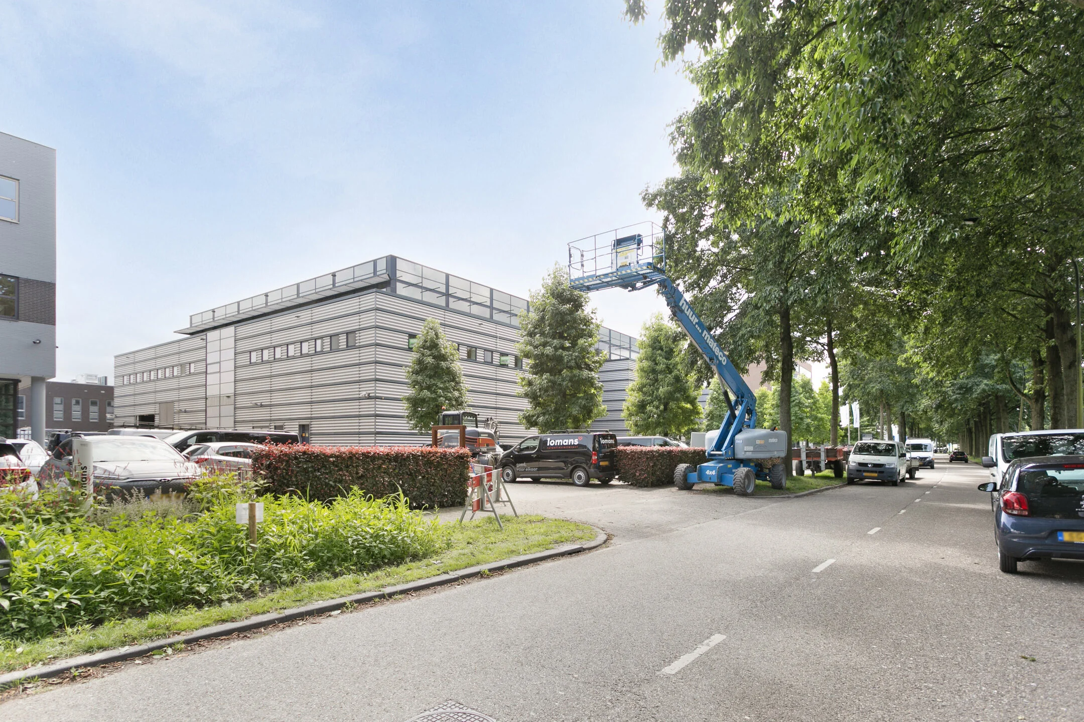 Een modern kantoorgebouw met een hoogwerker en geparkeerde voertuigen langs de straat.