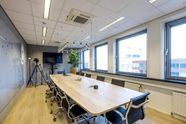 Een moderne vergaderruimte met een lange houten tafel, stoelen, een televisiescherm en grote ramen.