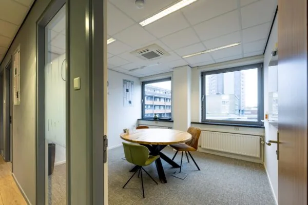 Een moderne vergaderruimte met een ronde houten tafel en stoelen bij grote ramen met uitzicht op omliggende gebouwen.