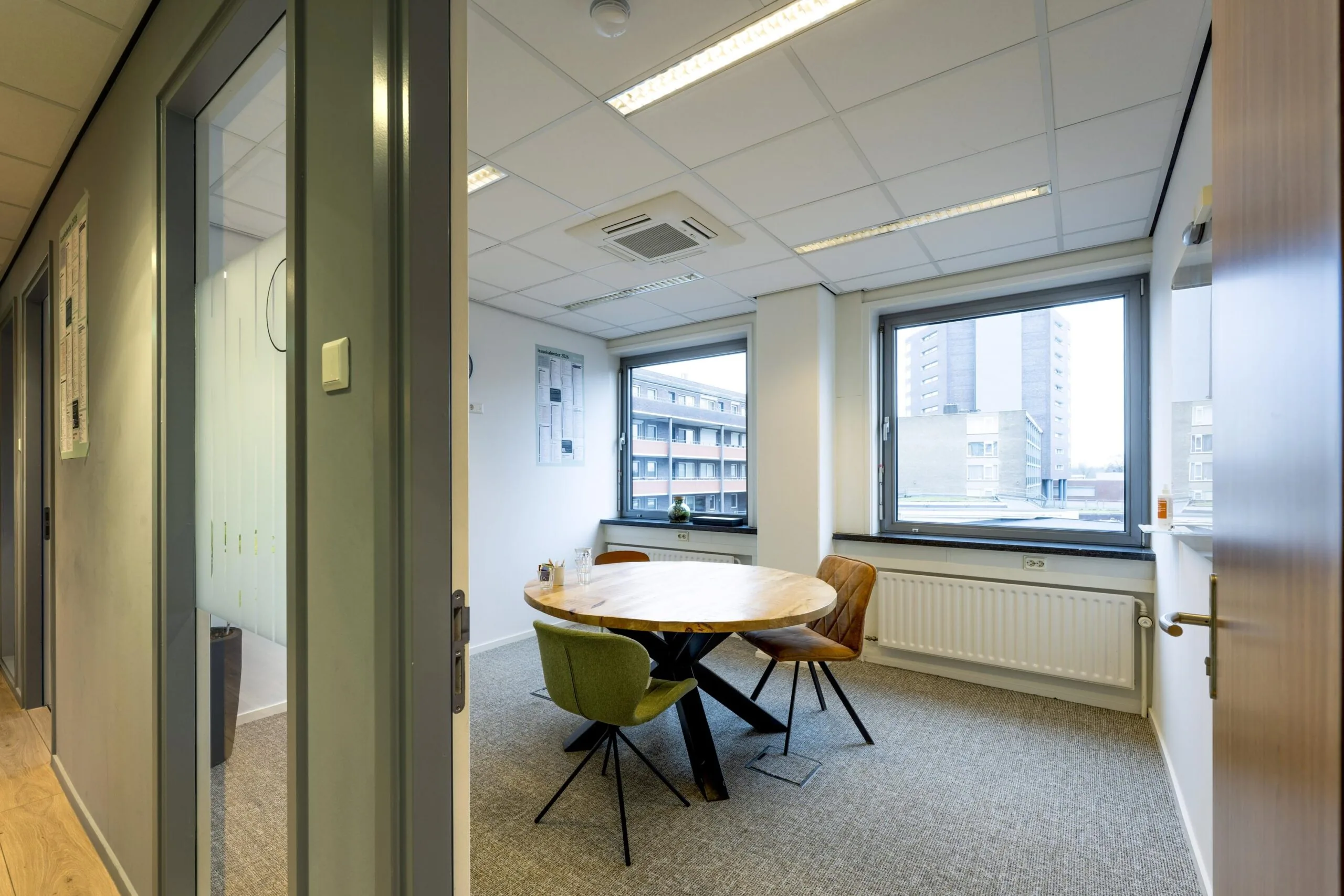 Een moderne vergaderruimte met een ronde houten tafel en stoelen bij grote ramen met uitzicht op omliggende gebouwen.