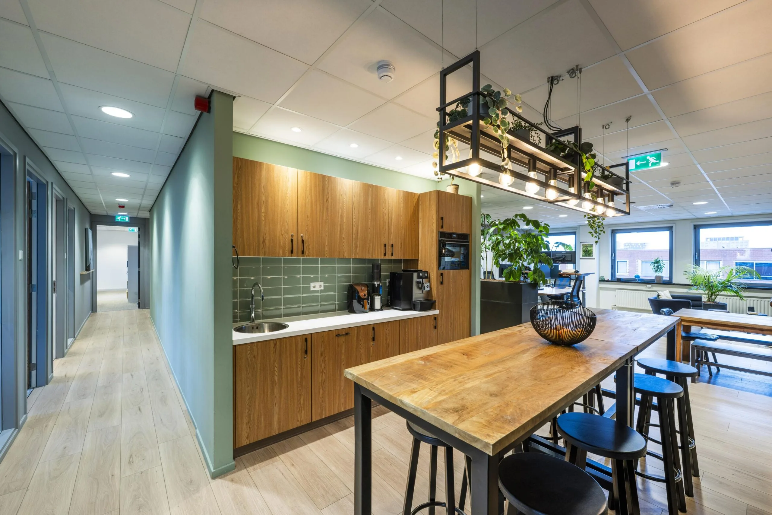 Moderne kantoorruimte met een keukenhoek en lange houten tafel omringd door hoge krukken, verlicht met hanglampen en decoratieve planten.