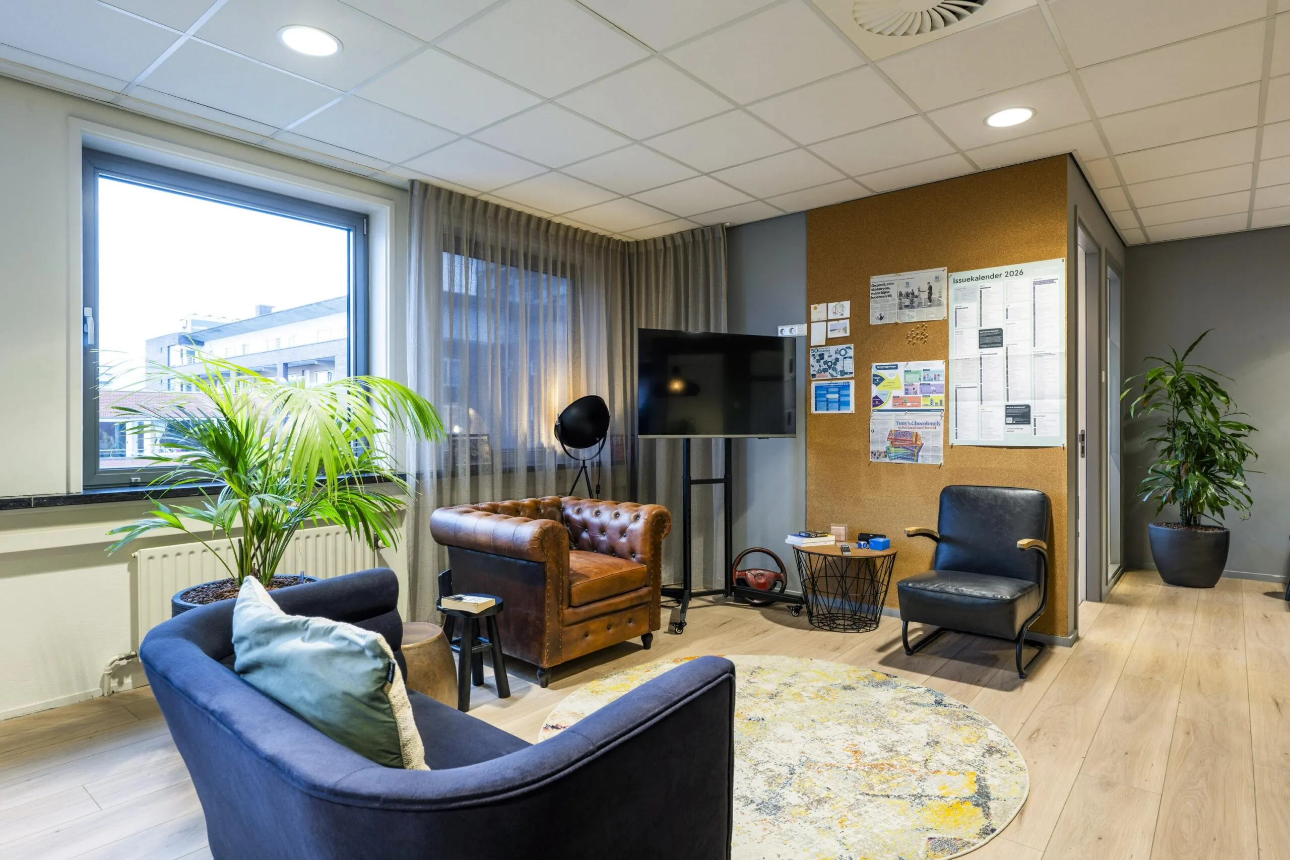 Een moderne kantoorruimte met diverse zitplaatsen, planten en een prikbord aan de muur.