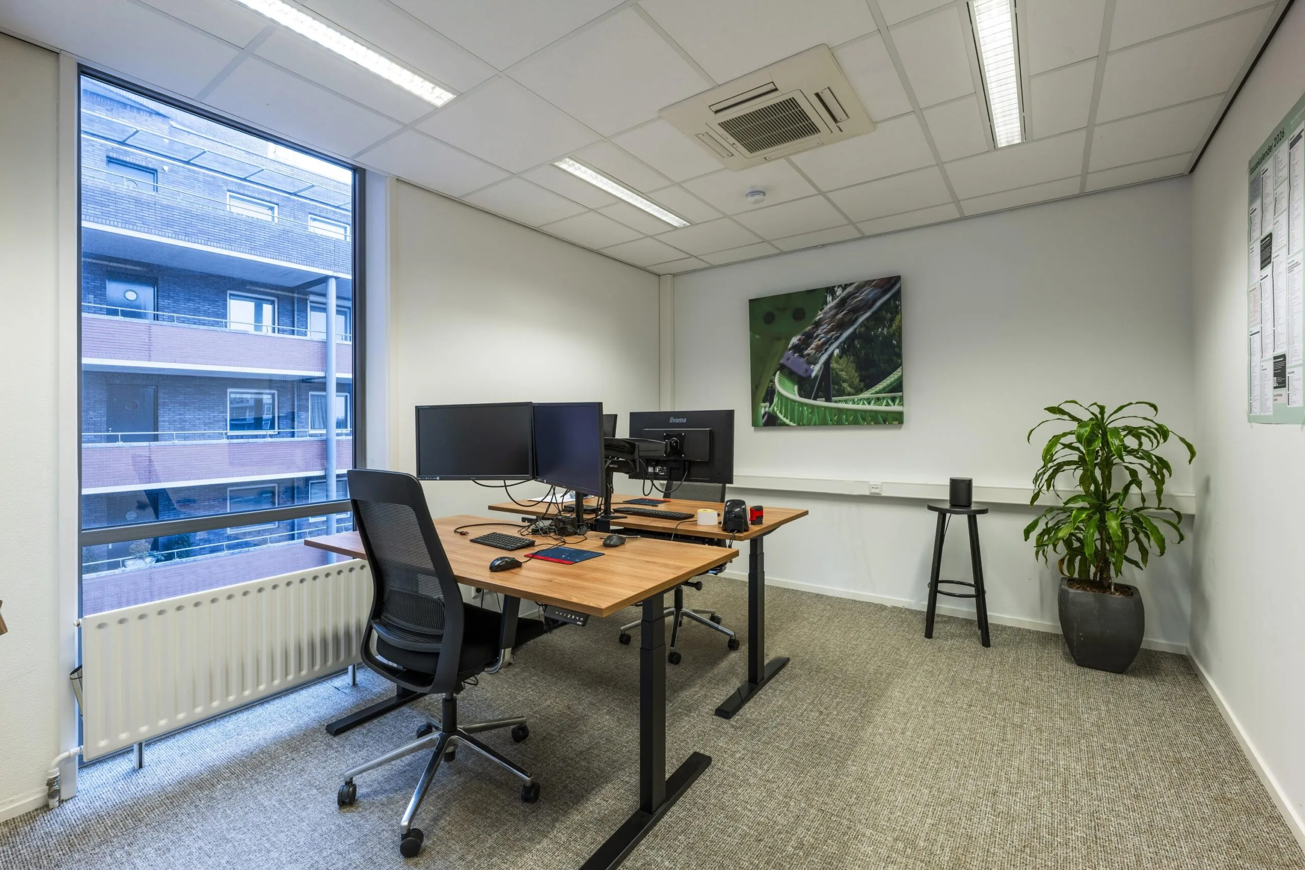 Een modern kantoor met een bureau, twee monitoren, een bureaustoel en een grote plantenbak bij het raam.