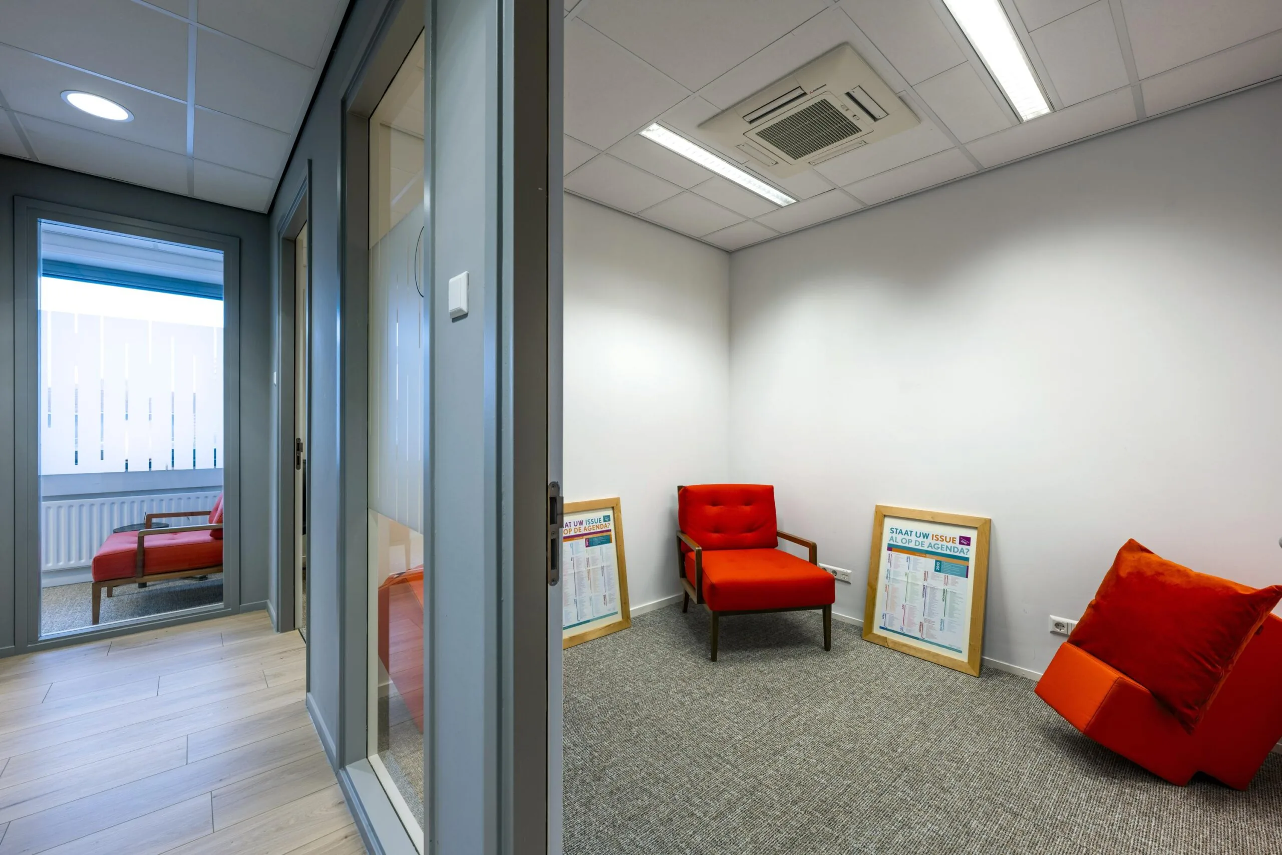 Een modern kantoor met een glazen wand toont een kamer met rode stoelen en ingelijste posters op de vloer.