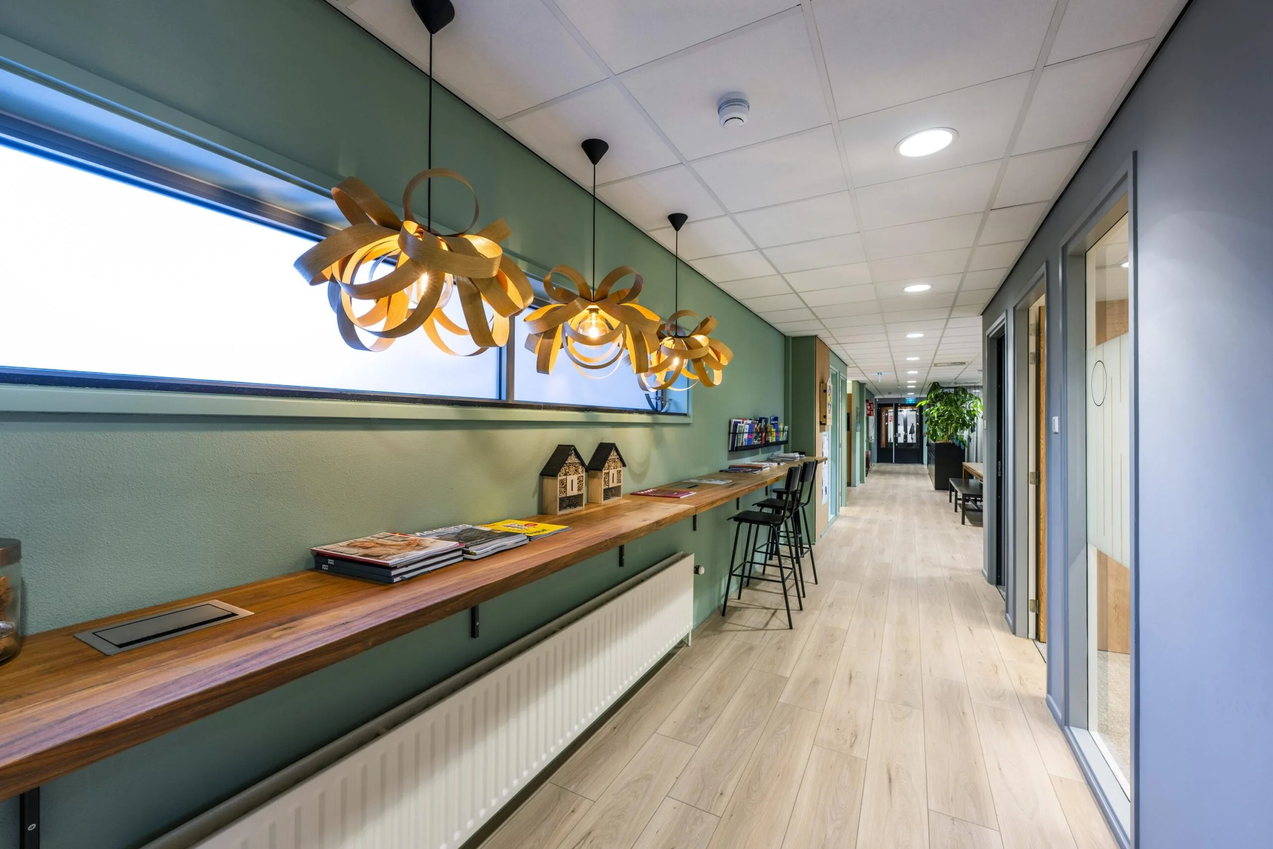 Een moderne gang met houten vloeren, hanglampen en een lange, houten plank met tijdschriften en krukken langs een groene muur.