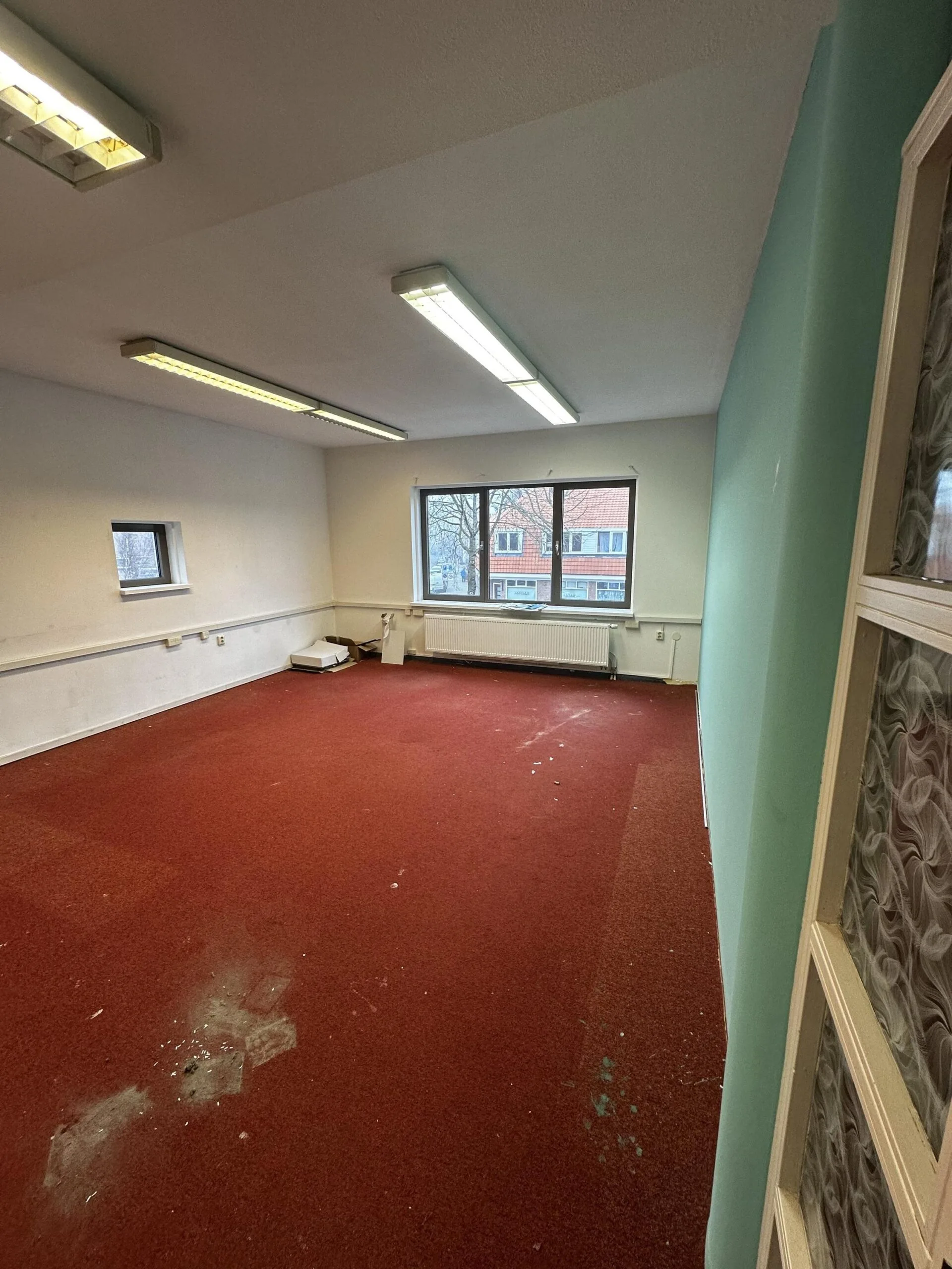 Leegstaande kamer met rode vloerbedekking en TL-verlichting aan het plafond.