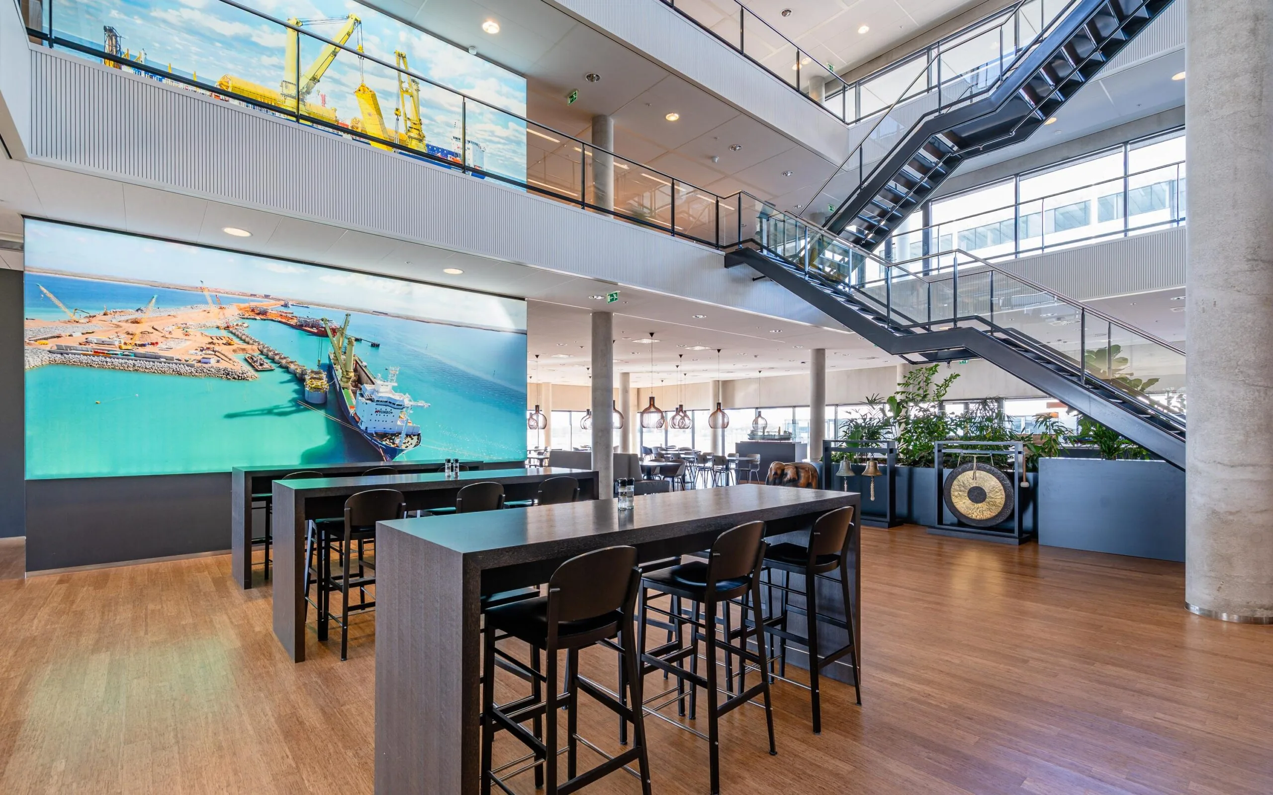 Moderne kantine met houten vloer en grote fotomuur van een haven, met tafels, stoelen, en een metalen trap.