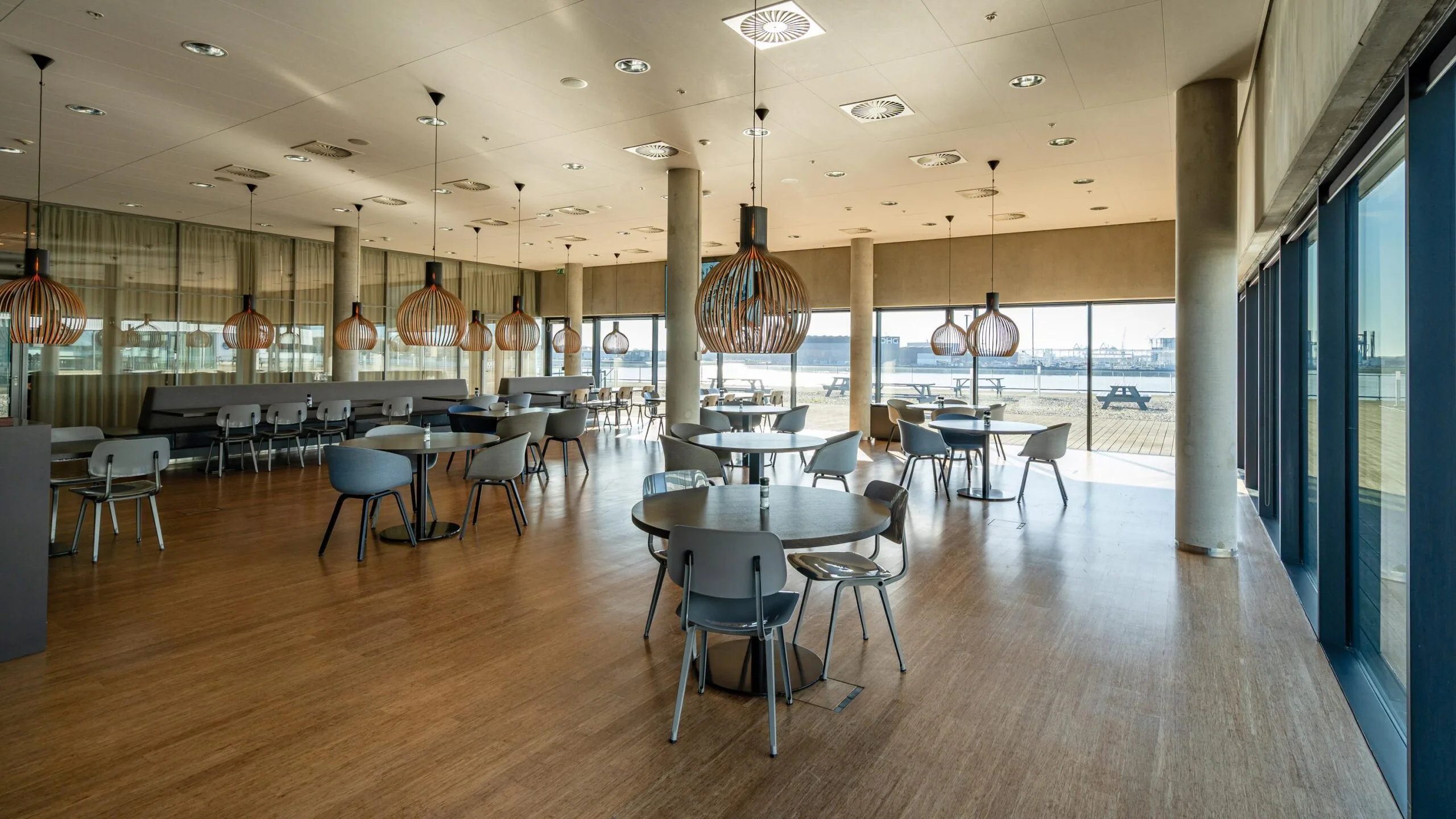 Een moderne, lichte eetzaal met houten vloeren, ronde tafels en hanglampen, uitkijkend op de haven vanuit grote ramen.