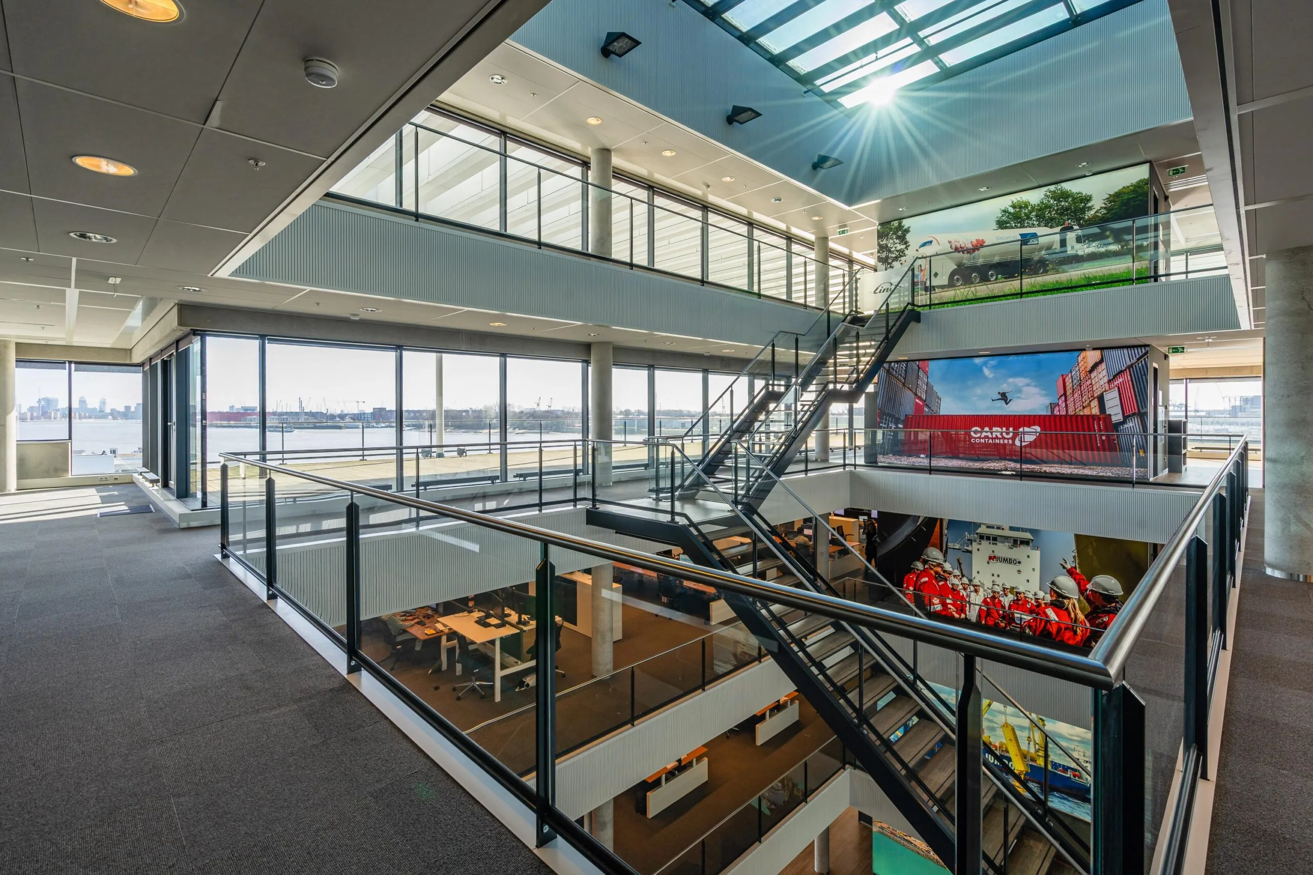 Interieur van een modern kantoor met open trappenhuis en grote ramen die uitzicht bieden op een haven.
