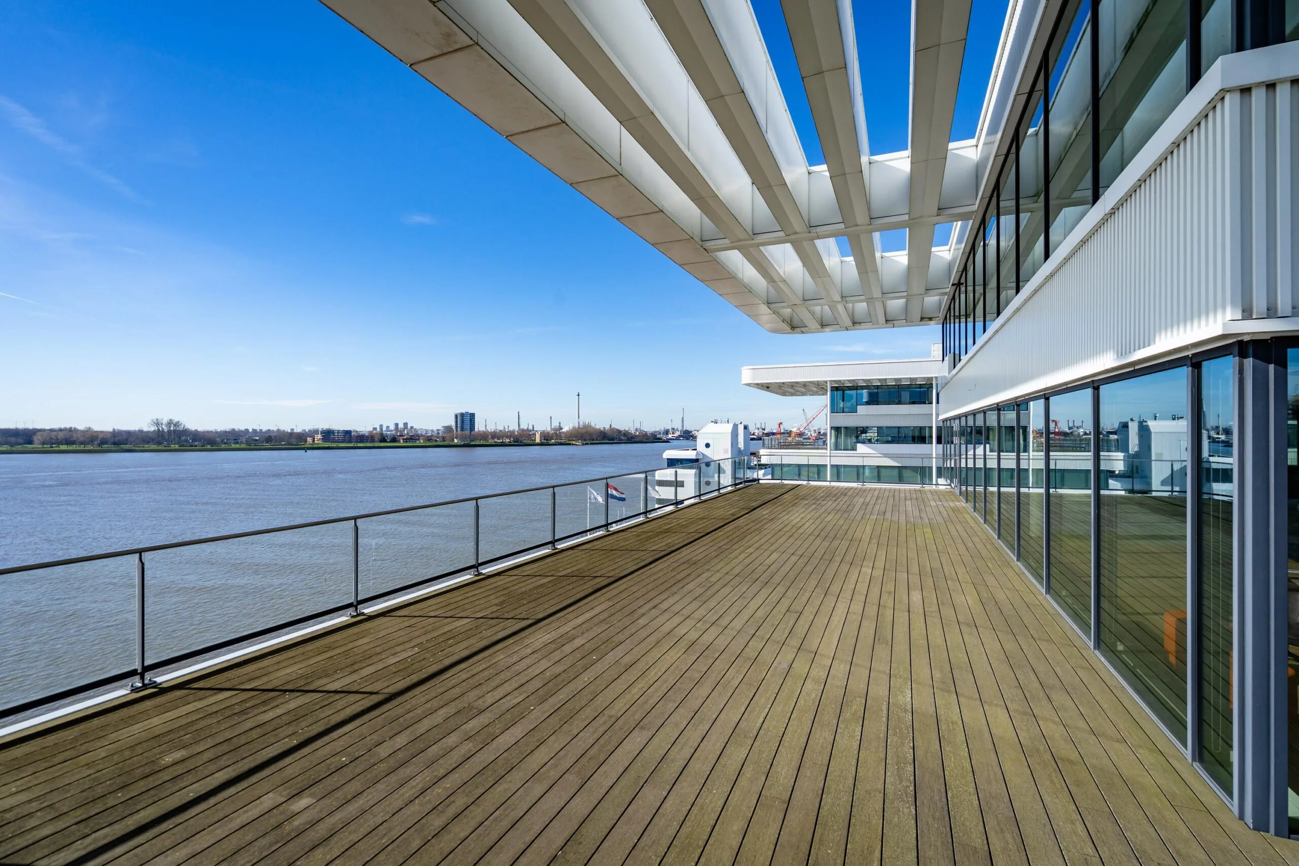 Uitzicht vanaf een houten terras met glazen balustrade, gelegen aan het water, met moderne gebouwen en een blauwe lucht.