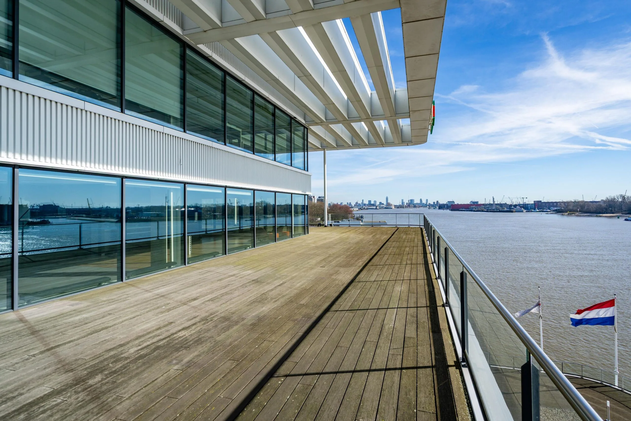 Modern gebouw aan de waterkant met een ruim, houten terras en uitzicht op de skyline, gefotografeerd bij helder weer.