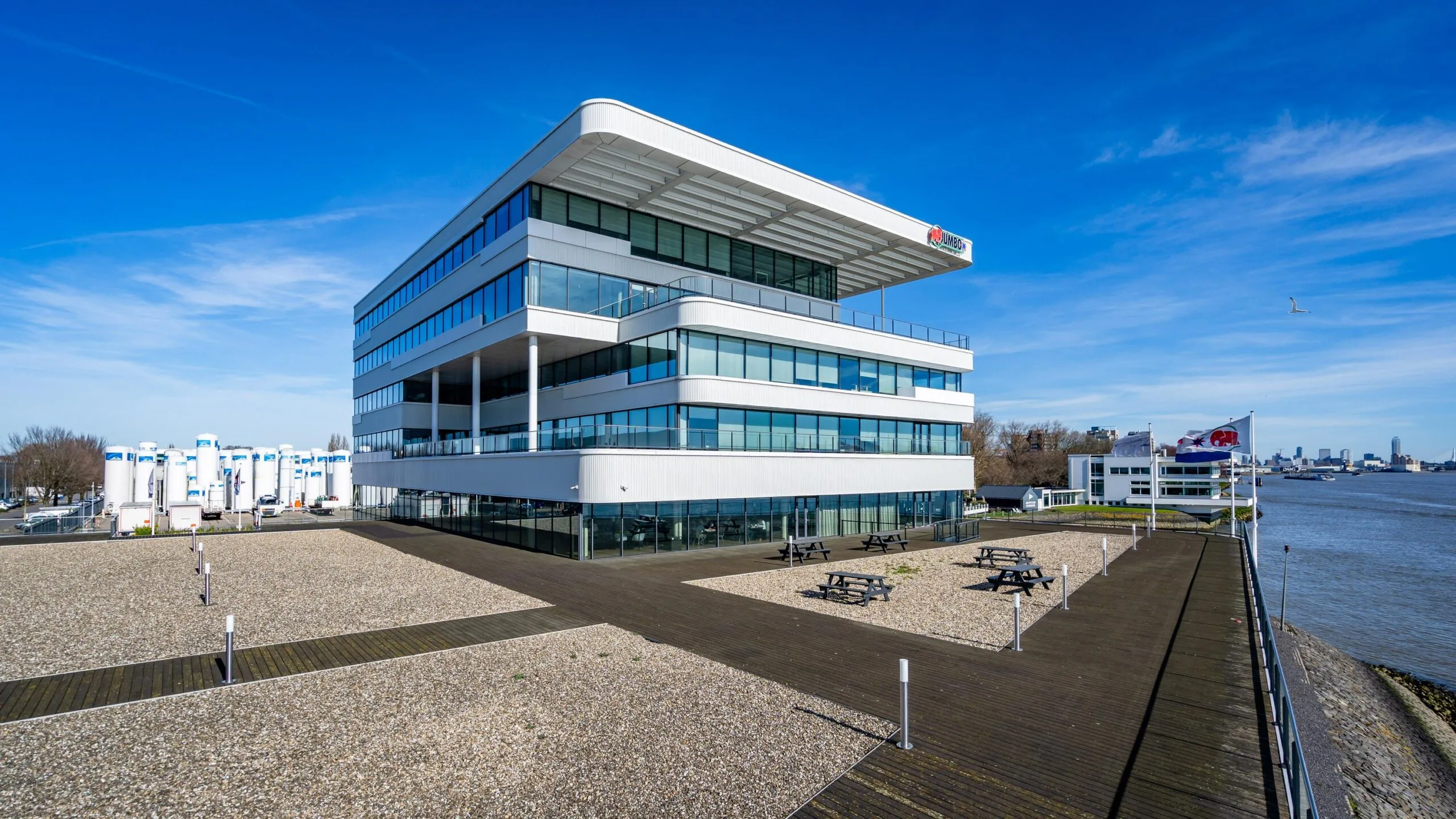 Modern, wit kantoorgebouw aan de rivier met blauwe lucht en wandelpaden.