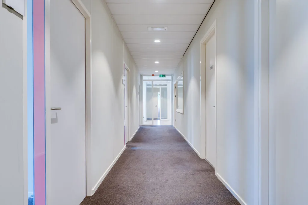 Een lange, smalle gang met wit geverfde muren, bruine vloerbedekking en meerdere deuren langs de zijkanten.