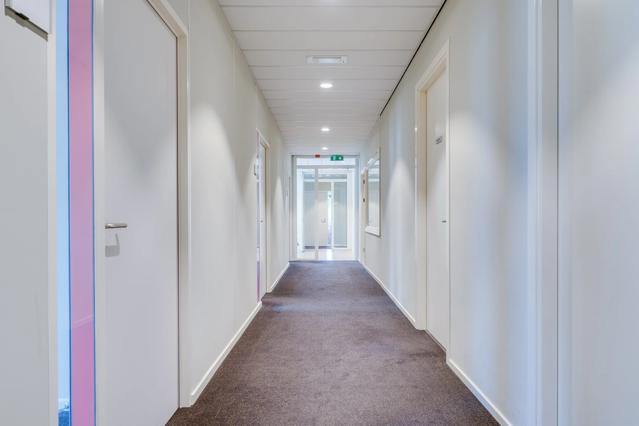 Een lange, smalle gang met wit geverfde muren, bruine vloerbedekking en meerdere deuren langs de zijkanten.