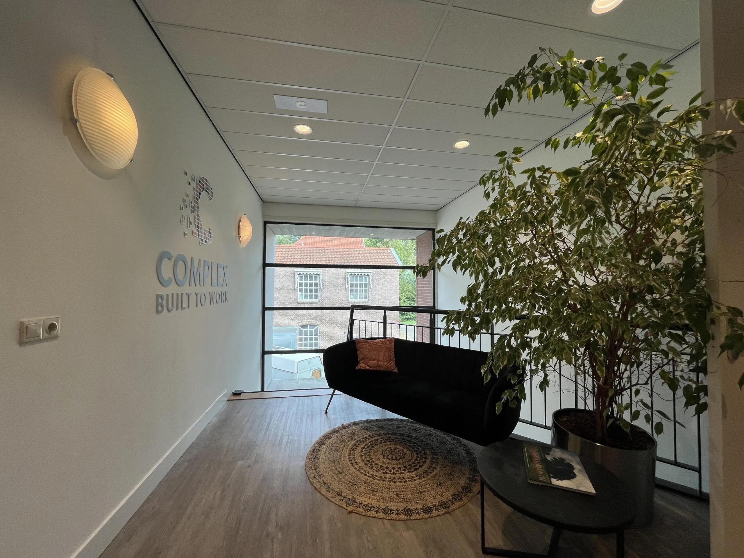 Moderne binnenruimte met een zwarte bank, grote plant, en raam met uitzicht op bakstenen gebouwen.