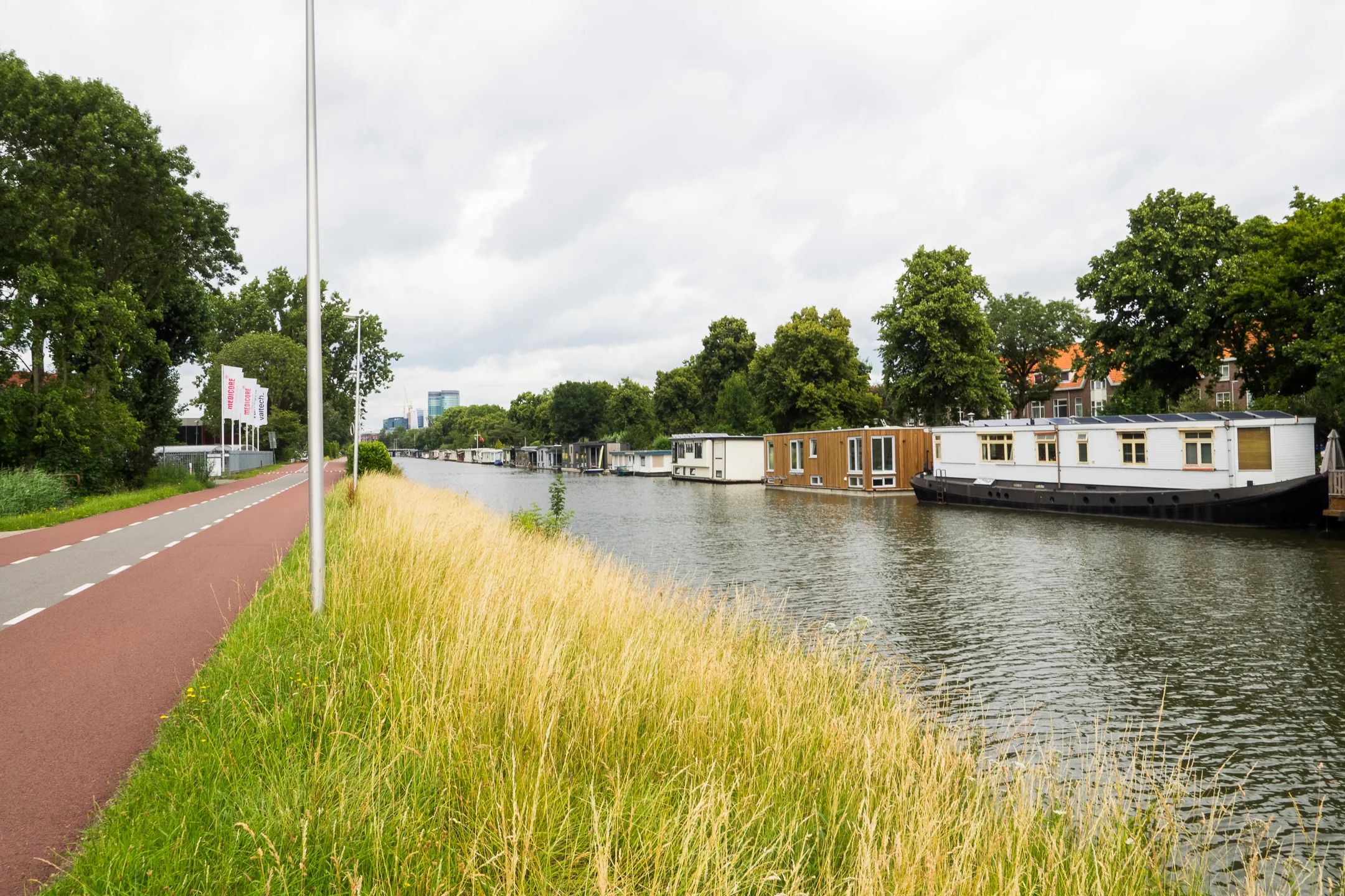 Fietspad naast een kanaal met woonboten en groene bomen op de achtergrond.