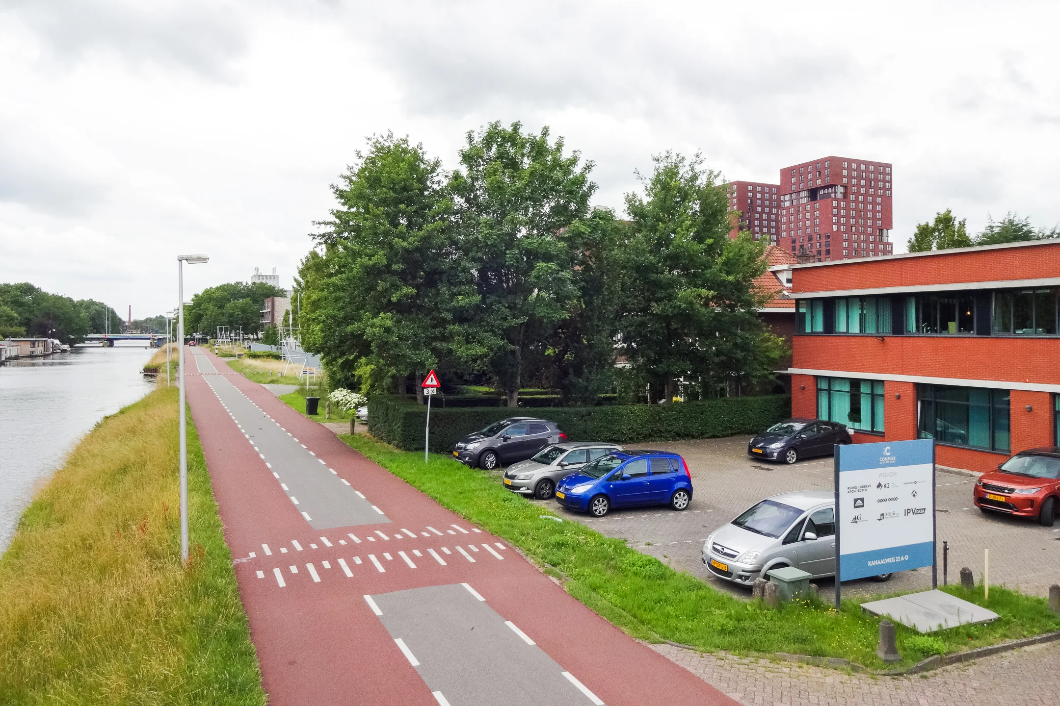Een fietspad langs een kanaal met geparkeerde auto's naast een rood bakstenen gebouw aan de Kanaalweg.