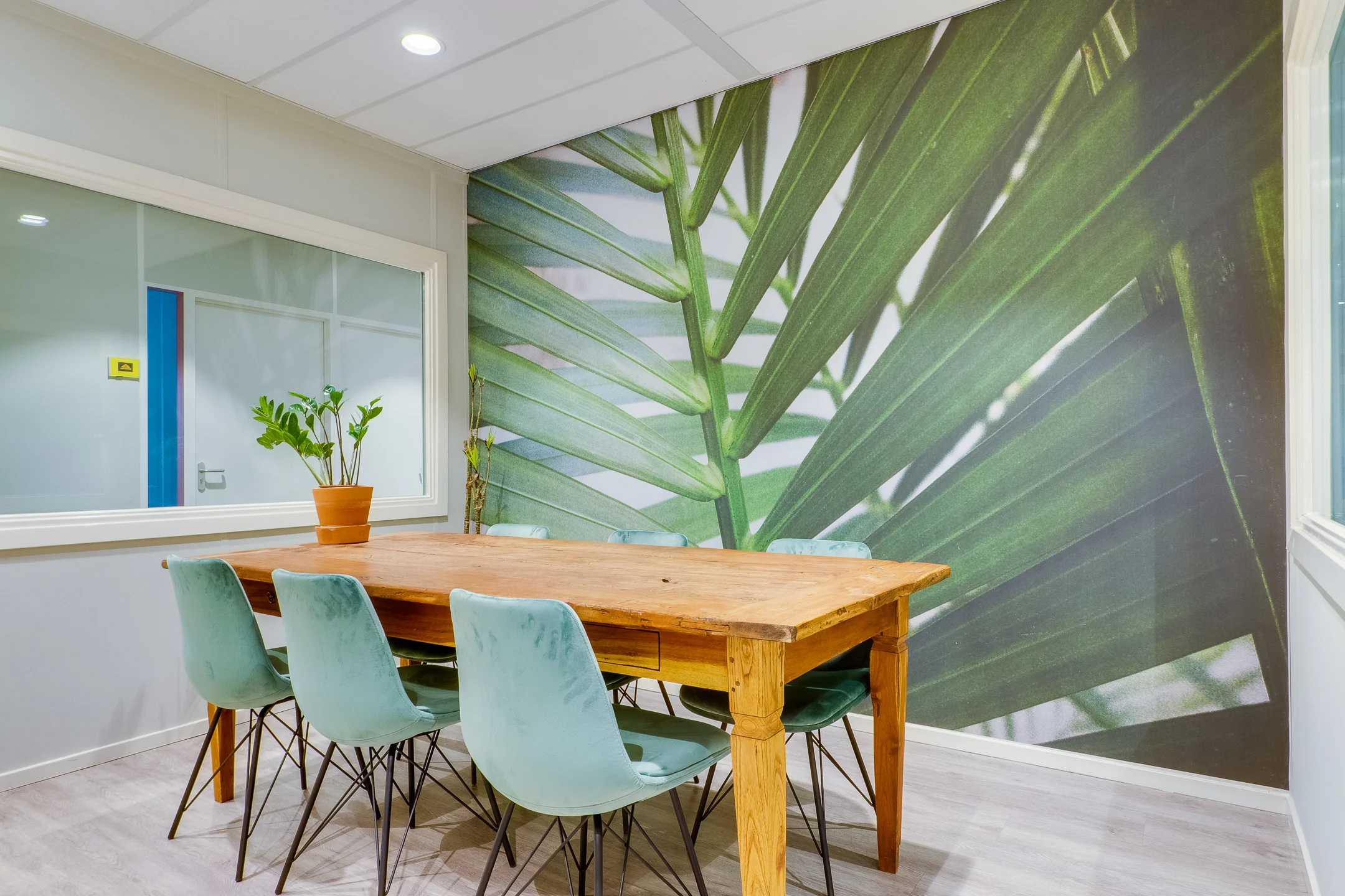 Een vergaderruimte met een houten tafel, groene stoelen en een muurdecoratie van palmbladeren.