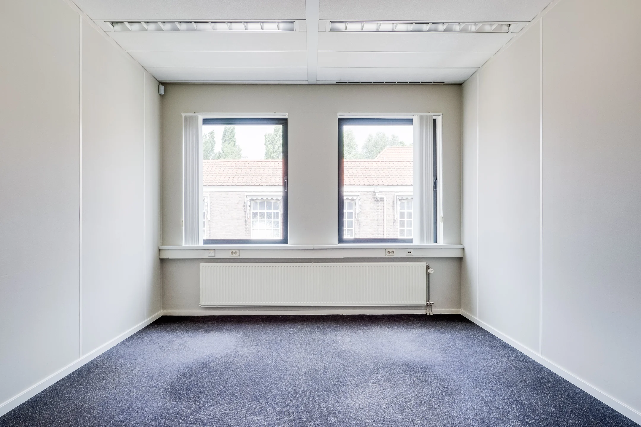 Een lege, lichte kamer met twee ramen en een radiator onder het raam.