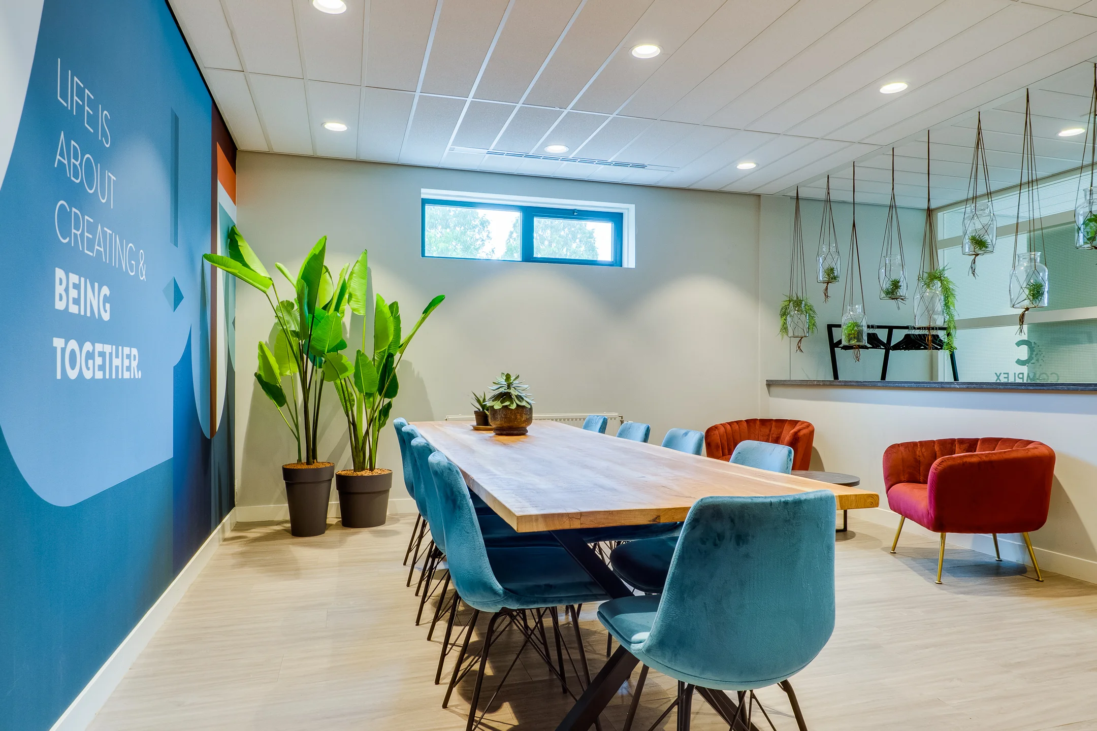 Een moderne vergaderruimte met een lange houten tafel, blauwe stoelen, kamerplanten en een inspirerende muurslogan.