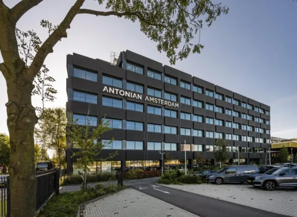 Een modern kantoorgebouw met de naam "Antonian Amsterdam" aan de Spaklerweg, omringd door bomen en geparkeerde auto's.