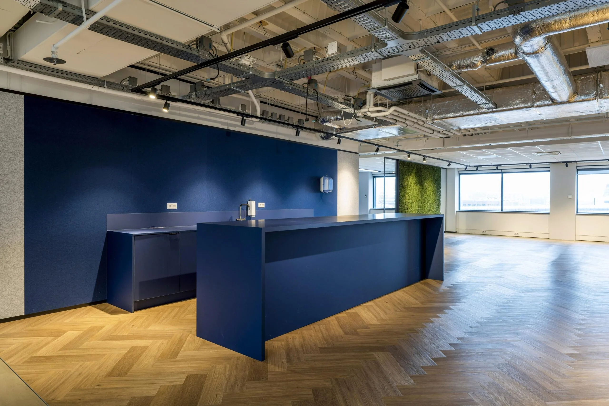 Een moderne kantoorruimte met een donkerblauwe balie, houten visgraatvloer en groene wand.