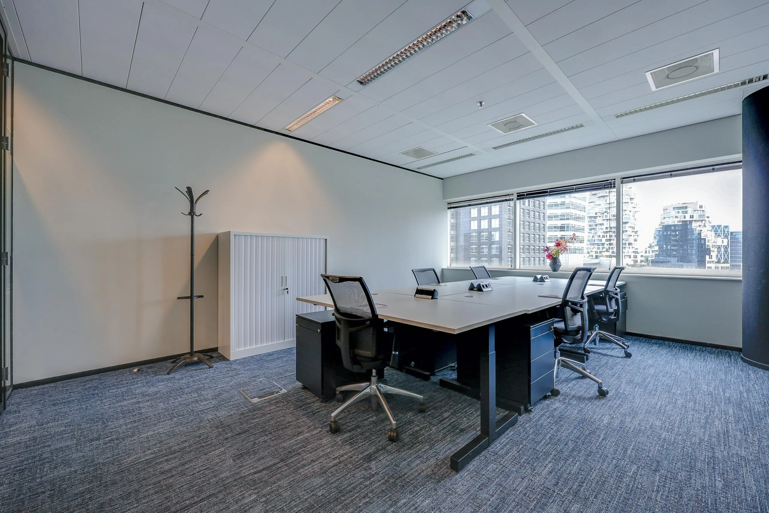 Een moderne kantoorruimte met bureaus en stoelen, omgeven door grote ramen die uitzicht bieden op gebouwen.