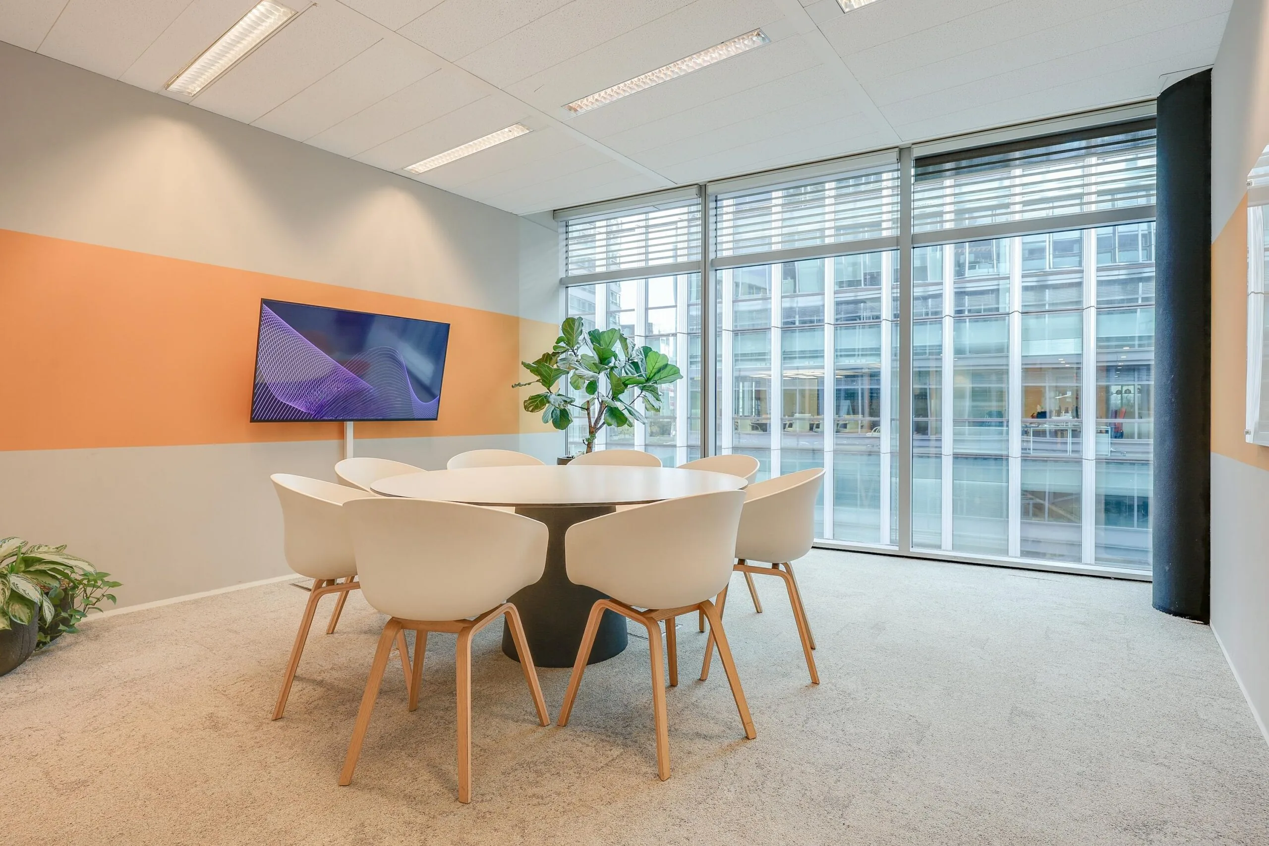 Vergaderruimte met een ronde tafel, witte stoelen, een groot raam en een scherm aan de muur.