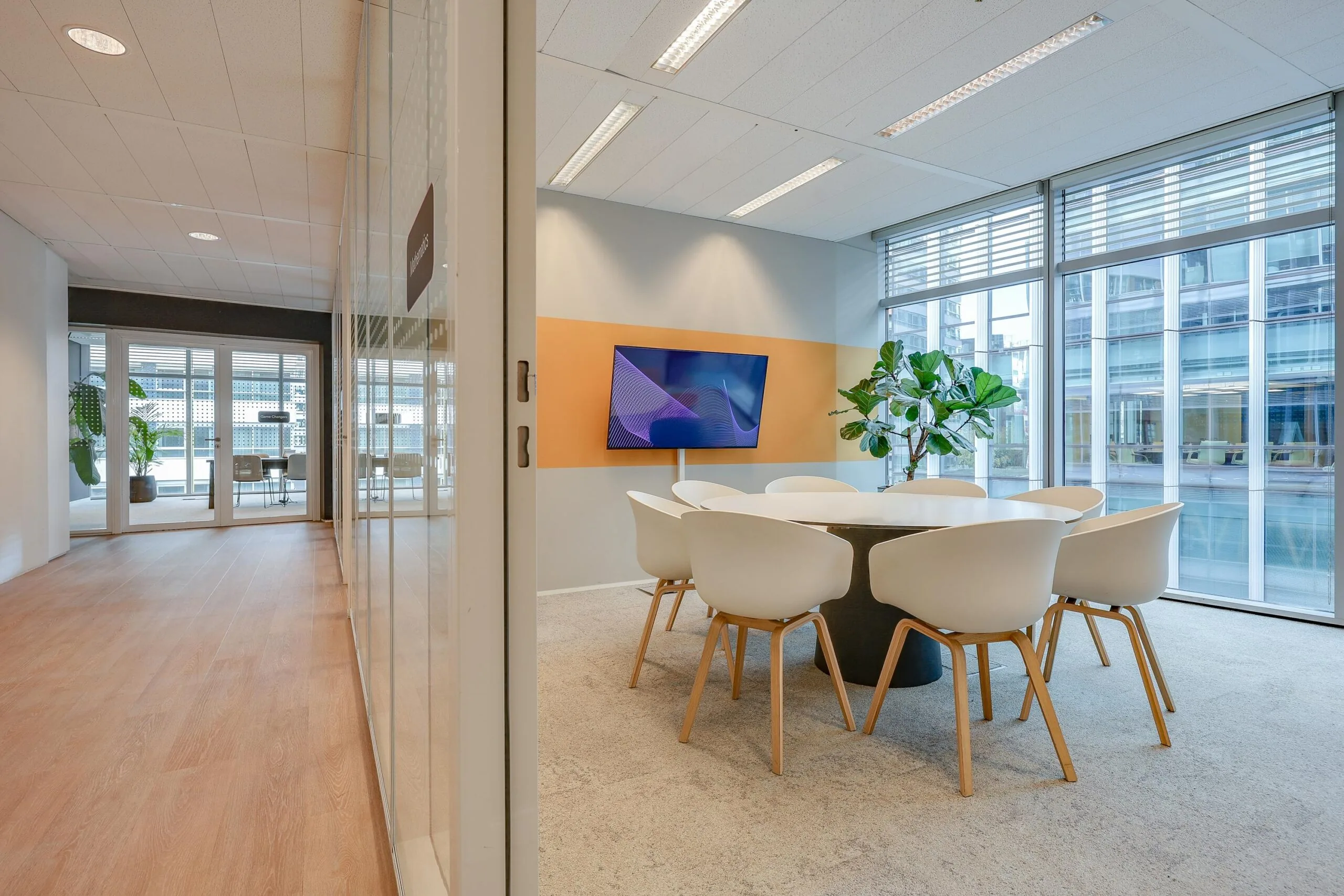 Een moderne vergaderruimte met een grote tafel, witte stoelen en een scherm, omgeven door glazen wanden.